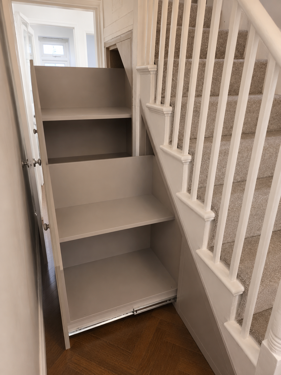 Empty built-in white storage shelves next to staircase with beige carpeted steps and white railing in a home interior.