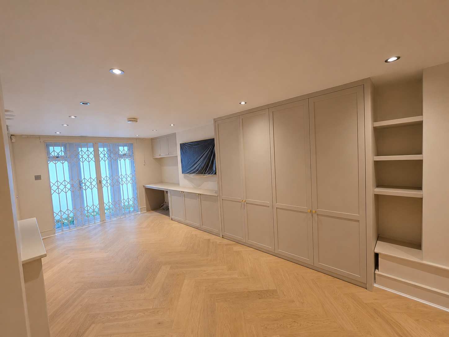 Empty living room with beige built-in cabinets, a television covered with plastic, hardwood floors, and sliding glass doors with geometric patterned security grille.