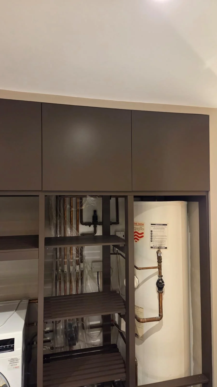Part of a laundry or utility room with dark brown cabinets, shelving, and copper piping against an off-white wall and ceiling.