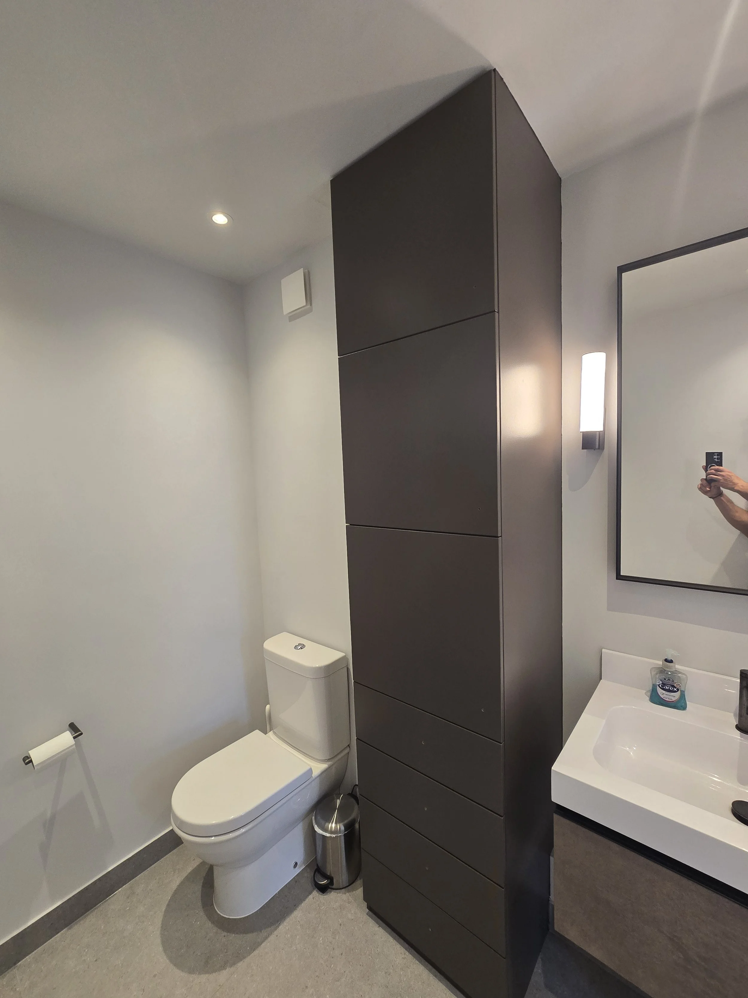 A modern bathroom with a white toilet, a gray floor, and a dark brown cabinet on the wall. There is a wall-mounted light fixture, a mirror, and a small white sink with a soap dispenser. The person taking the photo is partially visible in the mirror.