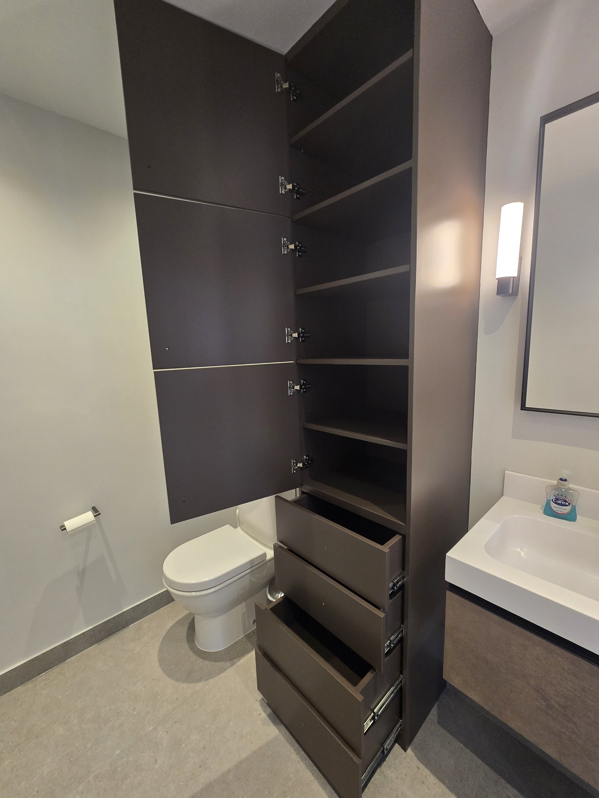 Empty bathroom with black wall-mounted storage cabinet, toilet, white sink with faucet, bottle of hand soap on sink, and wall-mounted light fixture.