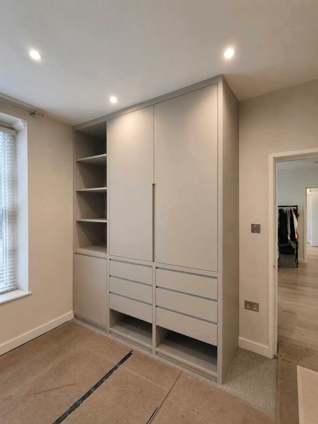 A built-in white closet with open shelves, drawers, and closed cabinet doors in a room under renovation, with partial view of an adjacent walk-in closet.
