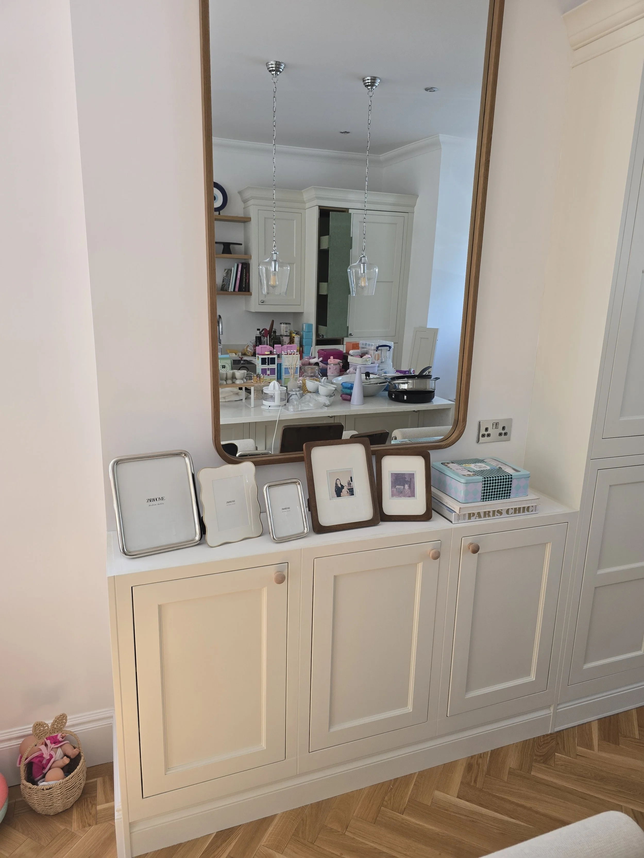 A large mirror above a white dresser with closed cabinets. The dresser has framed photographs, a decorative box, and various small items on top. Reflects a kitchen with white cabinets, hanging lights, and a cluttered countertop with kitchen supplies 