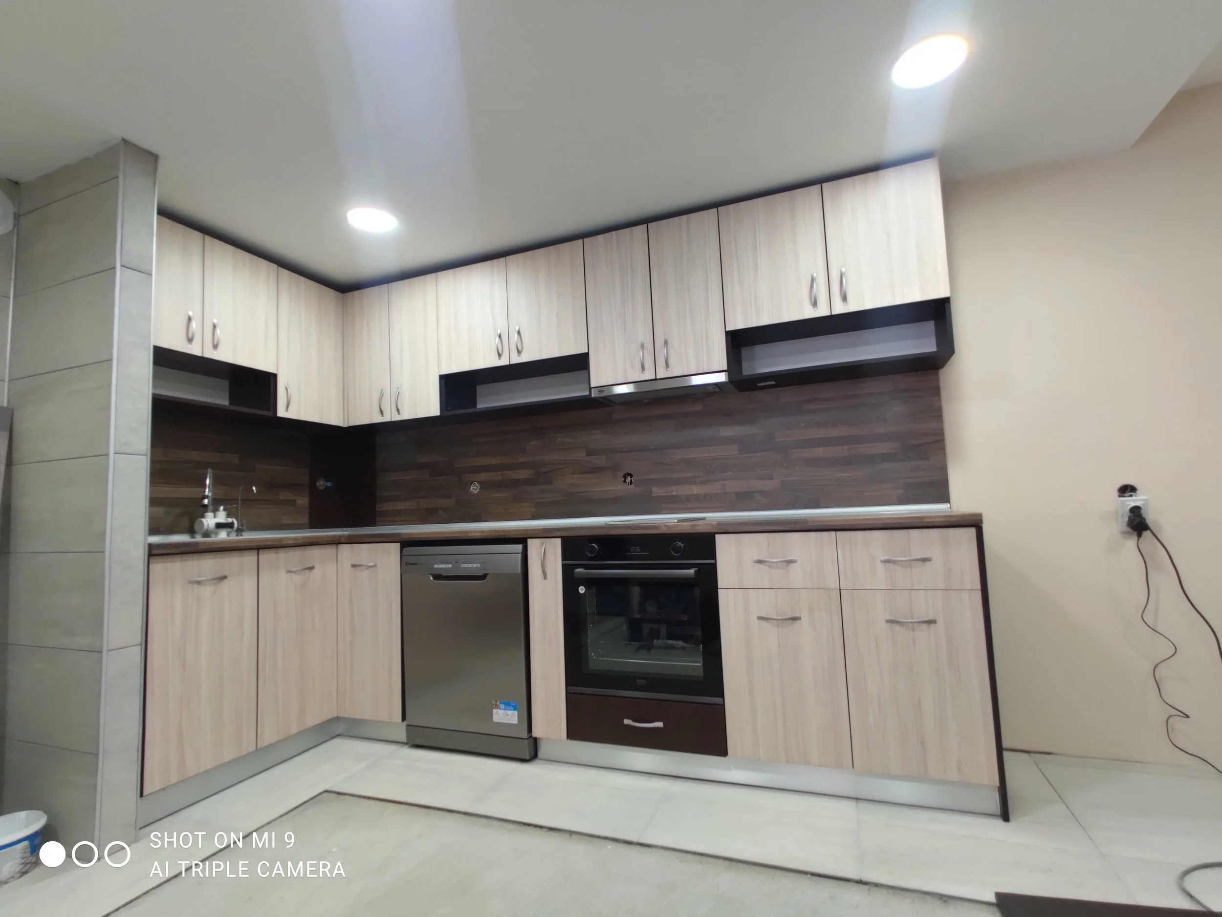 Kitchen with light wood cabinets, dark backsplash, built-in oven, dishwasher, and stovetop, with a tiled floor and electrical outlets on the wall.