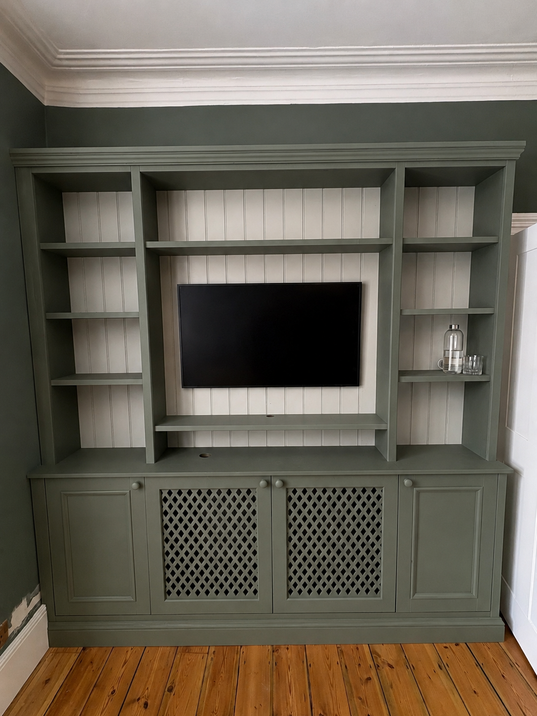 Green painted built-in bookshelf and cabinet unit with a mounted flat-screen TV in the center, white beadboard backing, and a small collection of glass water bottles and glasses on the right shelf, set on a wooden floor with green walls and white cro