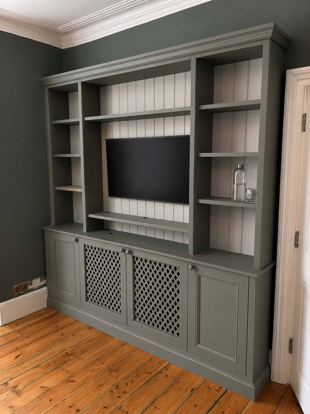 Gray built-in shelving unit with a television, decorated with a glass water bottle and glass on the right shelf, and a wooden floor in front.