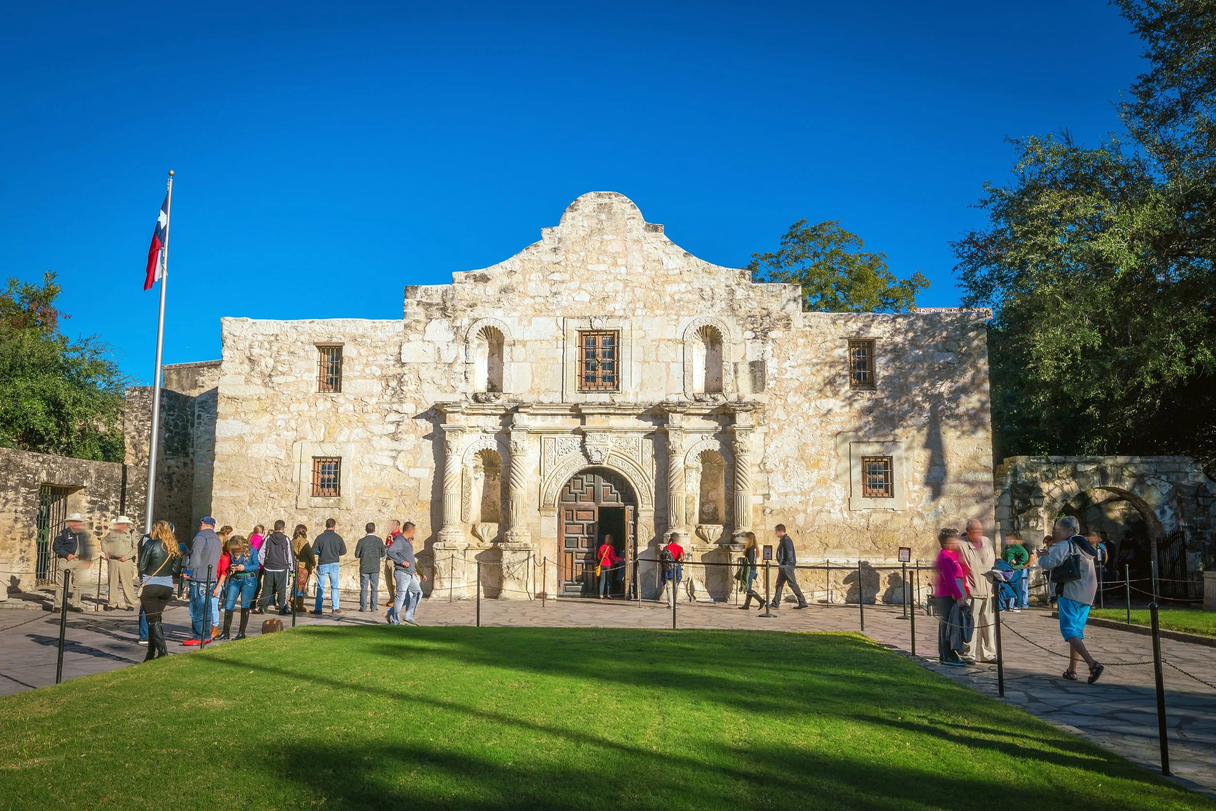 The Alamo, a historic white stone building with an ornate entrance, is surrounded by visitors and trees, with a Texas flag flying nearby under a clear blue sky.
