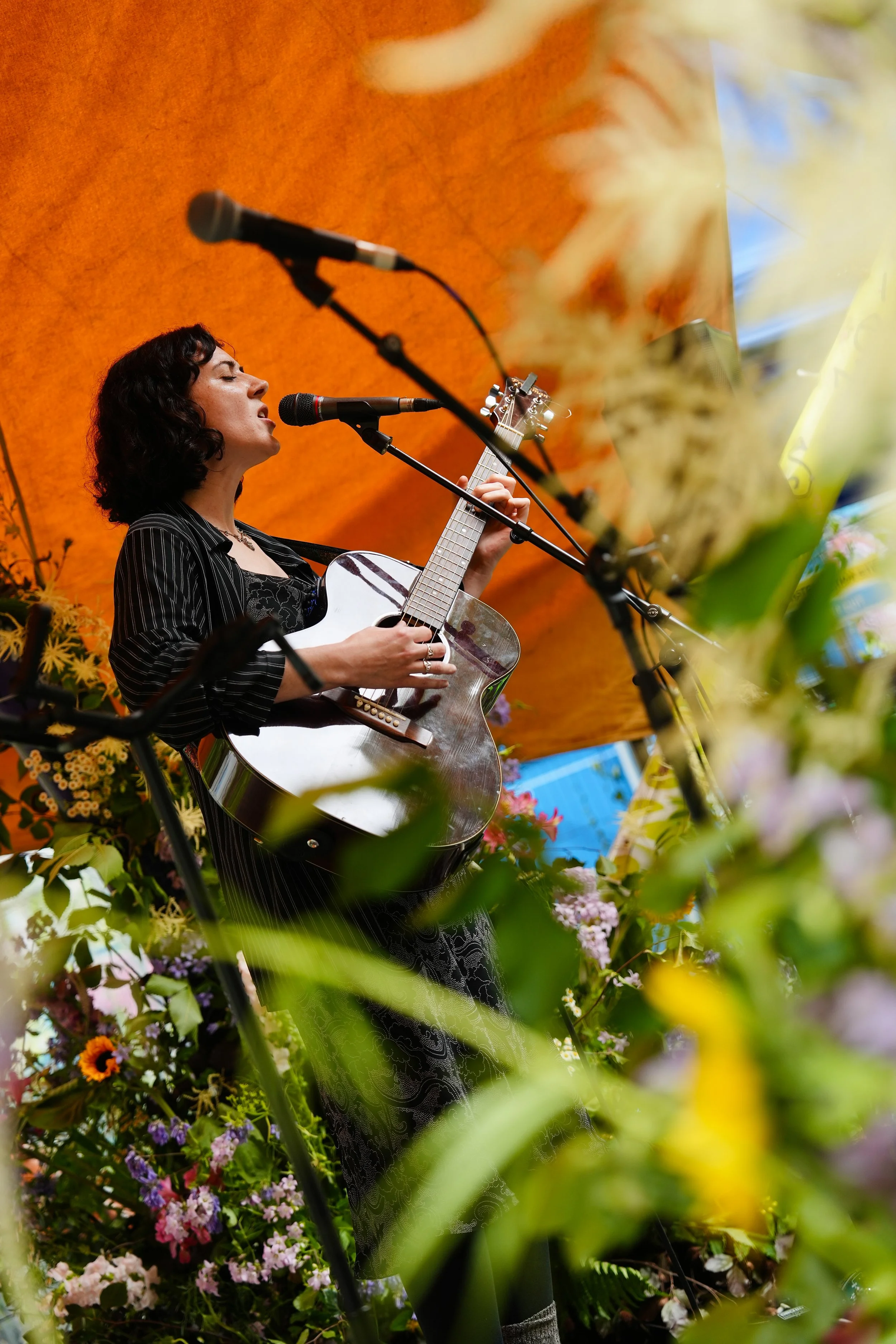A woman with dark, curly hair singing into a microphone and playing an acoustic guitar, surrounded by colorful flowers with an orange background.