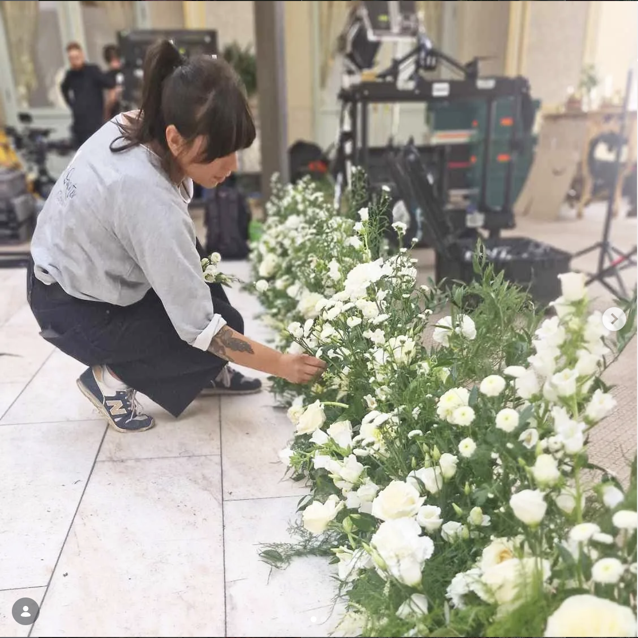 A woman with dark hair tied back, wearing a gray shirt and black pants, crouching down and arranging white flowers on a tiled floor, in an indoor setting. There is video equipment and other people in the background.