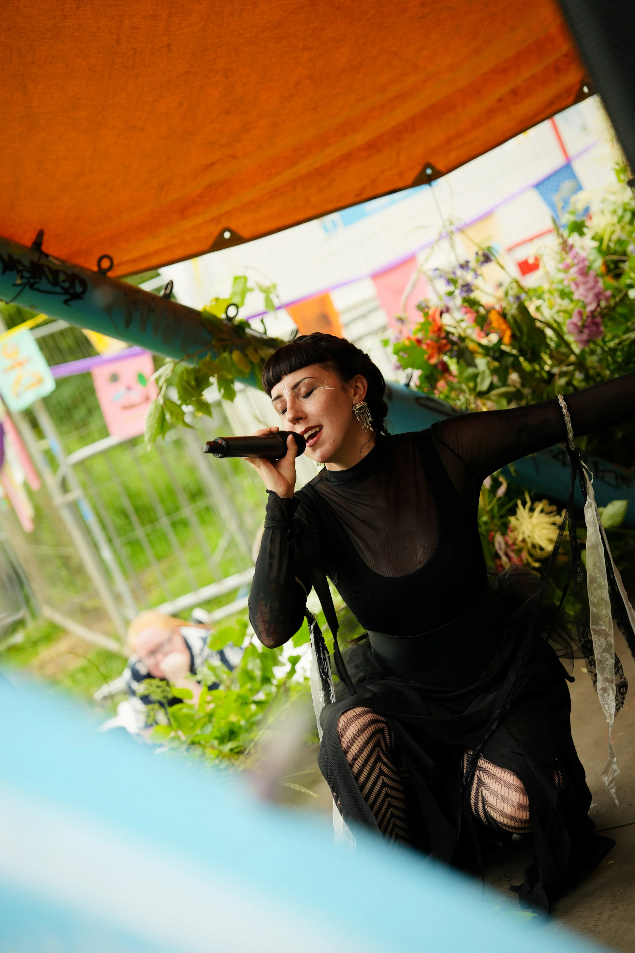 A woman with dark hair and makeup singing into a microphone at a colorful outdoor event with flowers and banners.