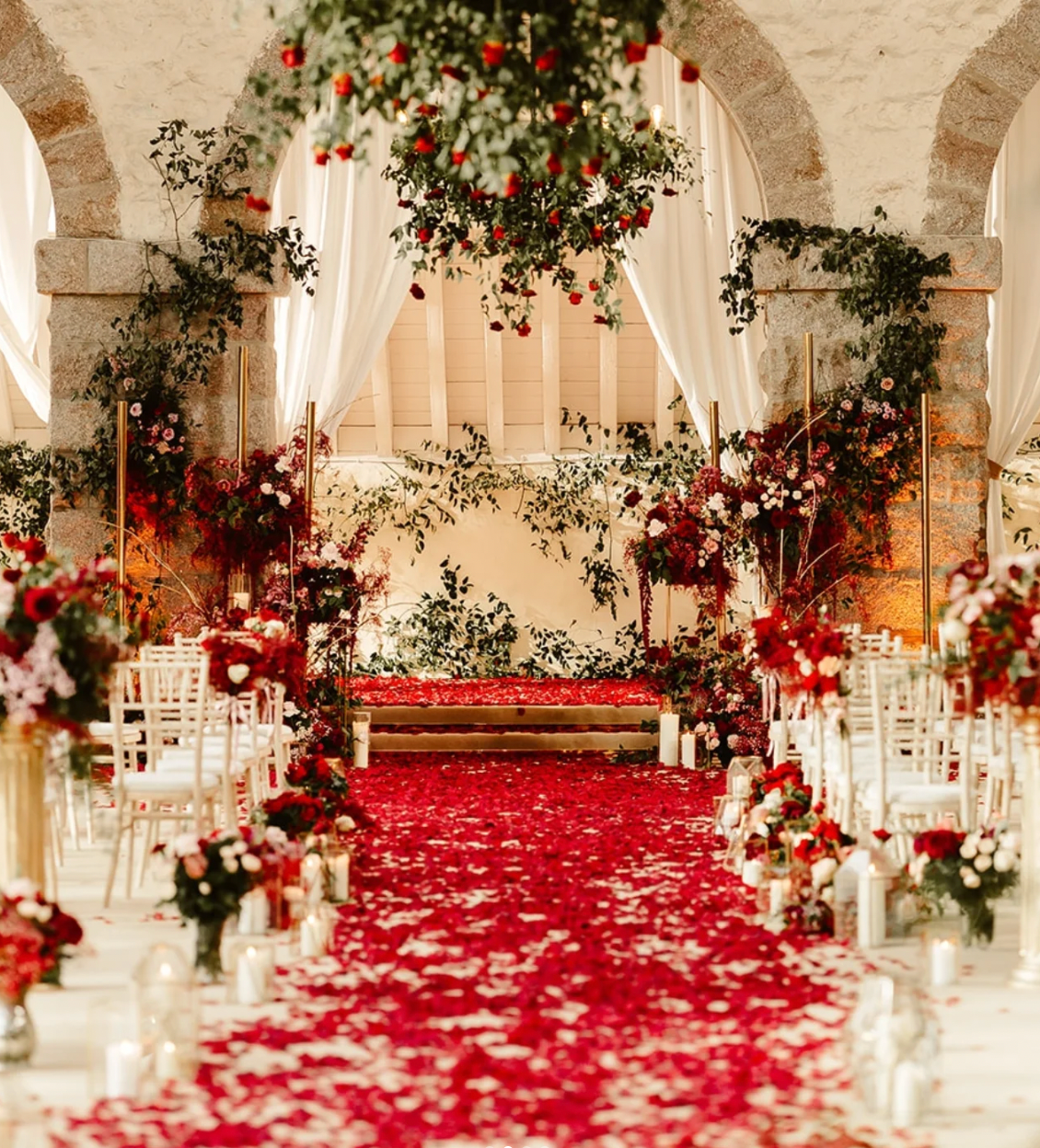 A romantic wedding ceremony setup with a red rose petal aisle, floral arrangements, candles, and elegant drapery in a stone-walled venue.