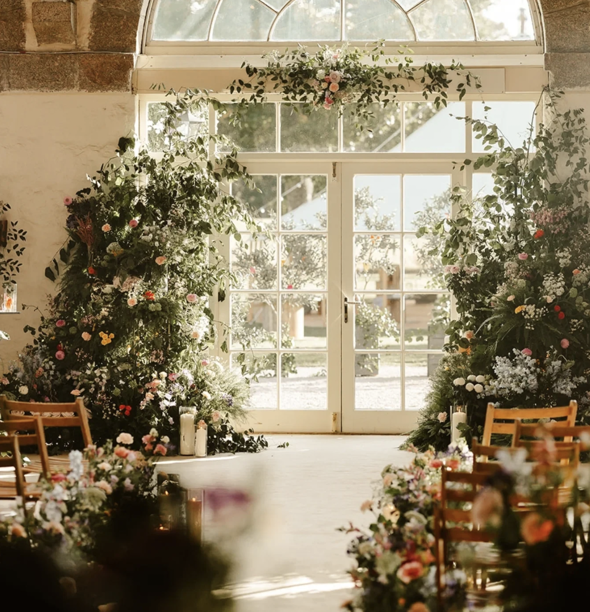 Beautiful indoor wedding or event space decorated with abundant flowers and greenery, with large glass doors and windows at the front letting in natural light.