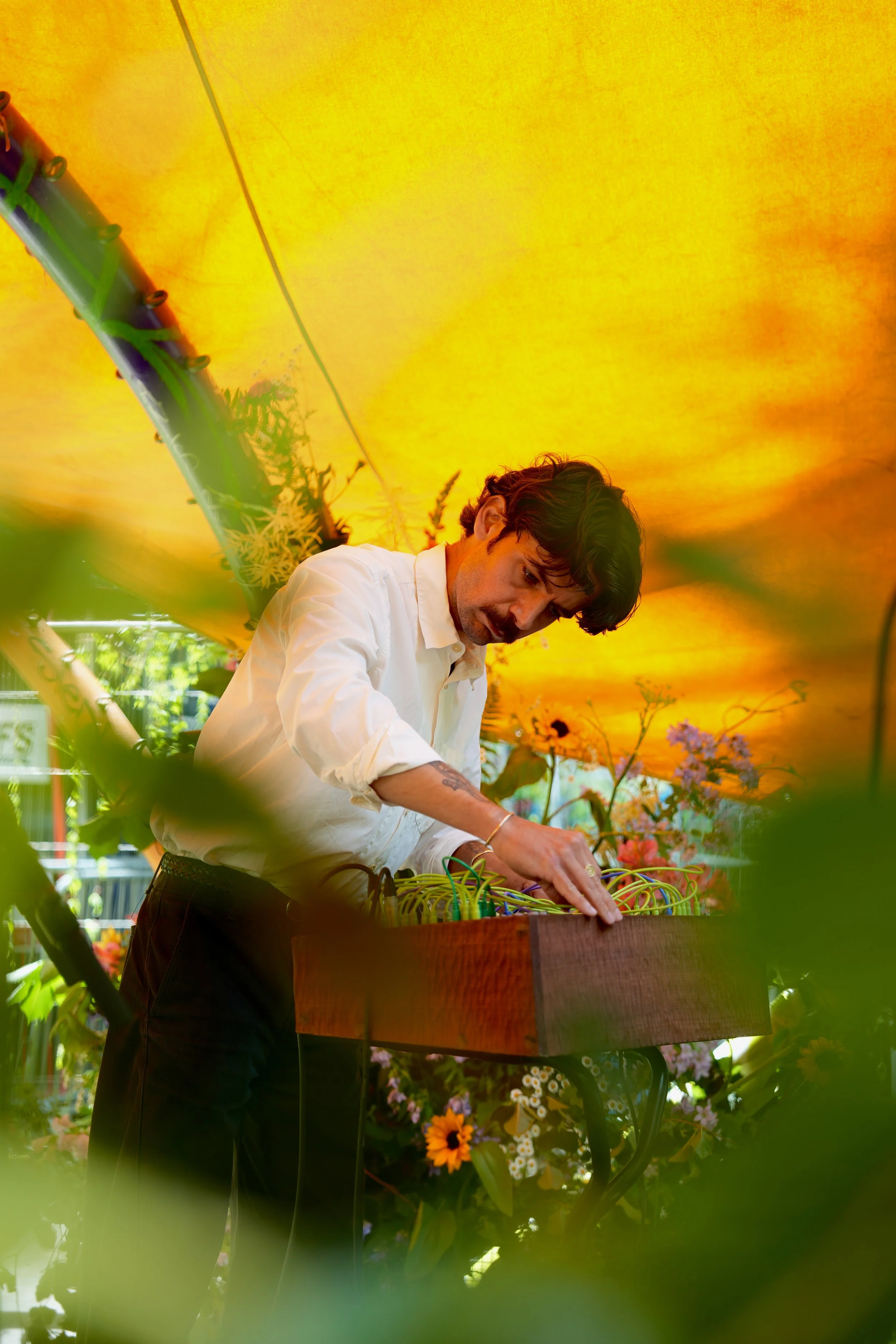 A man operating electronic music equipment at an outdoor event decorated with flowers, under a yellow canopy.