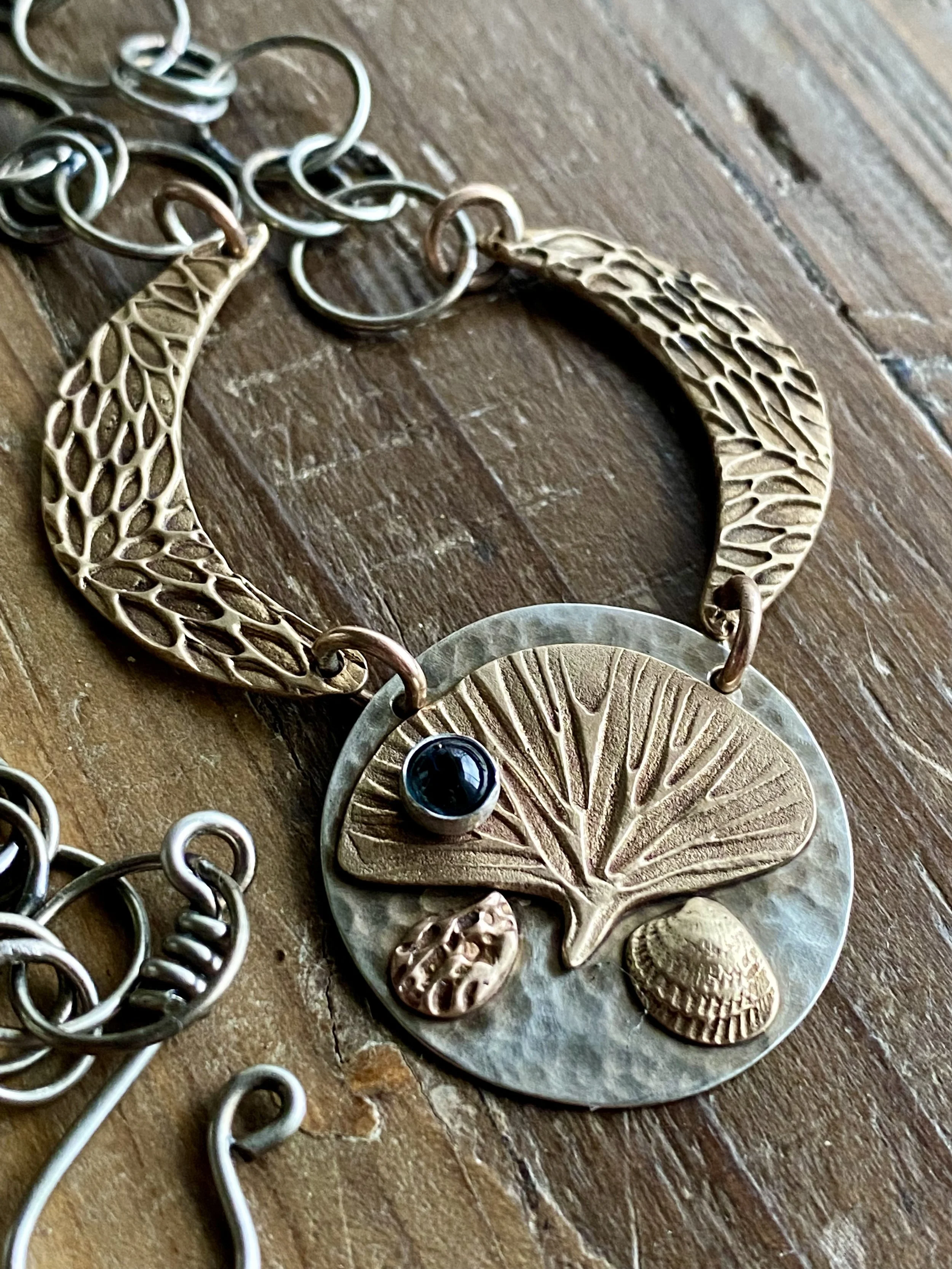 Close-up of various metallic jewelry pieces, including a leaf-shaped pendant with a blue gemstone and other circular and oval metallic decorations, placed on a wooden surface.