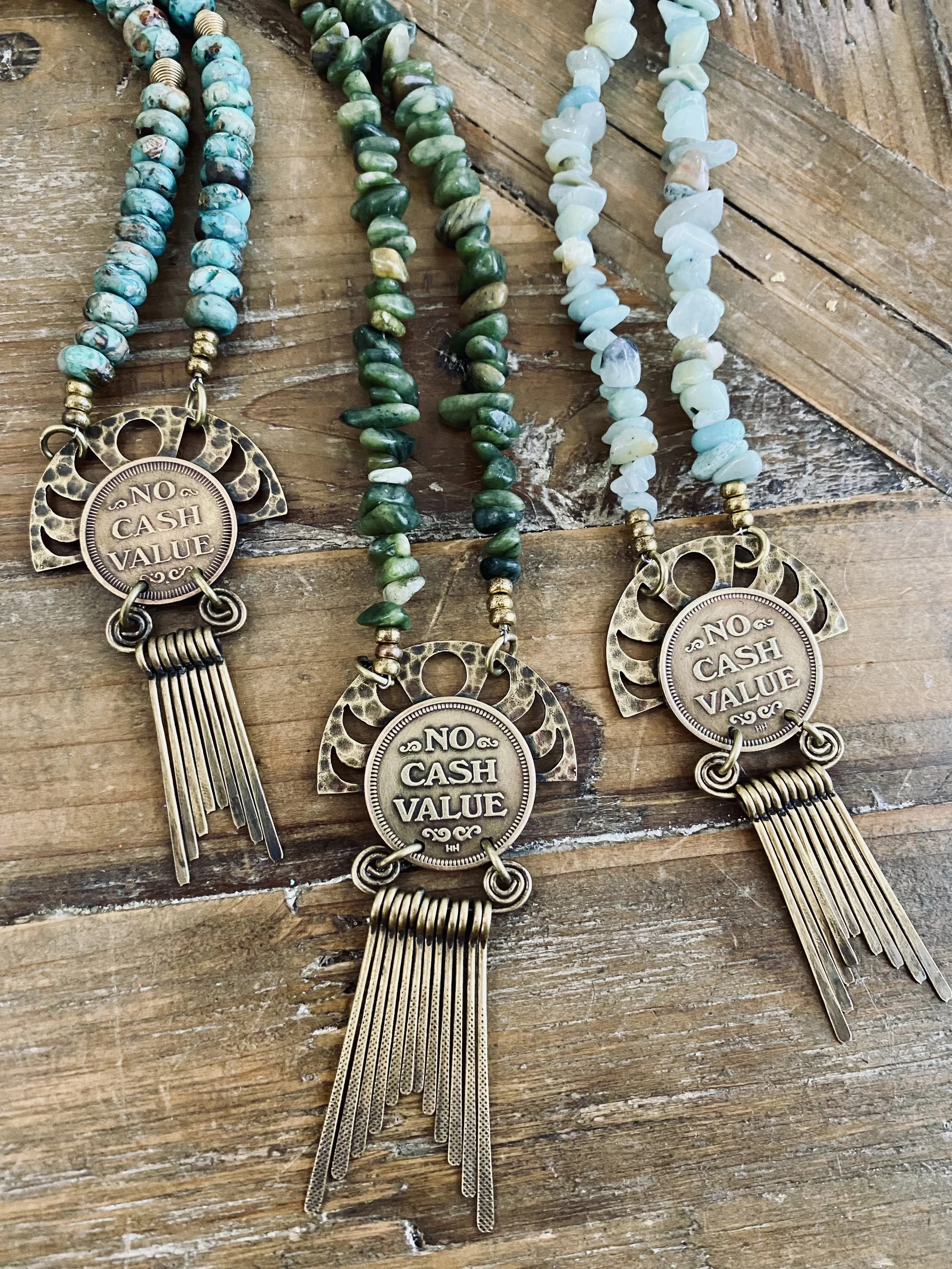 Four beaded necklaces with large metallic pendants that have the words 'NO CASH VALUE' engraved, lying on a wooden surface. 