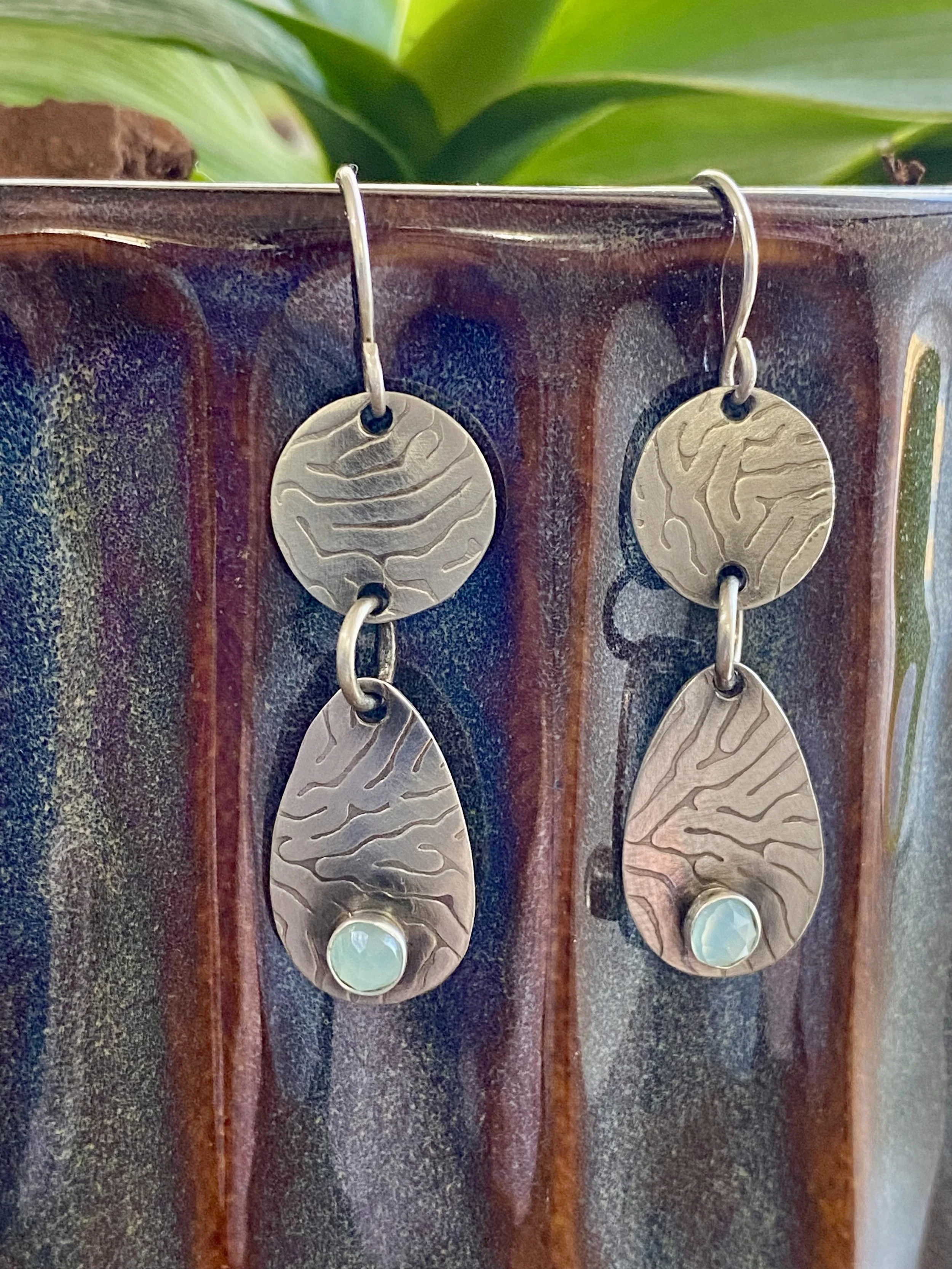 Close-up of a pair of earrings with circular, textured metal discs and iridescent stones at the bottom, hanging from a hook on a ceramic surface with green foliage in the background.