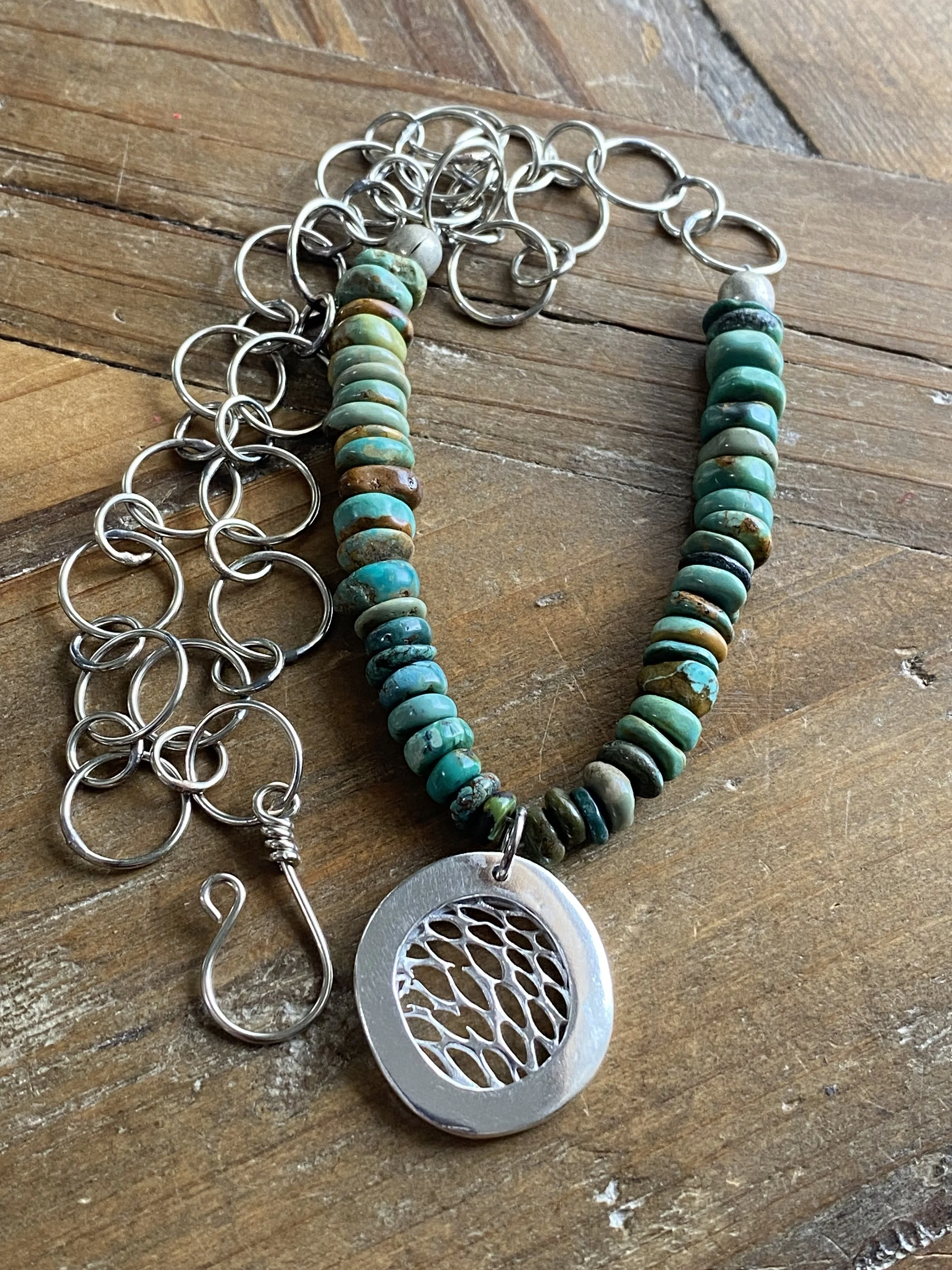 Metal necklace with turquoise beads and a circular pendant with a tree design, lying on a wooden surface.