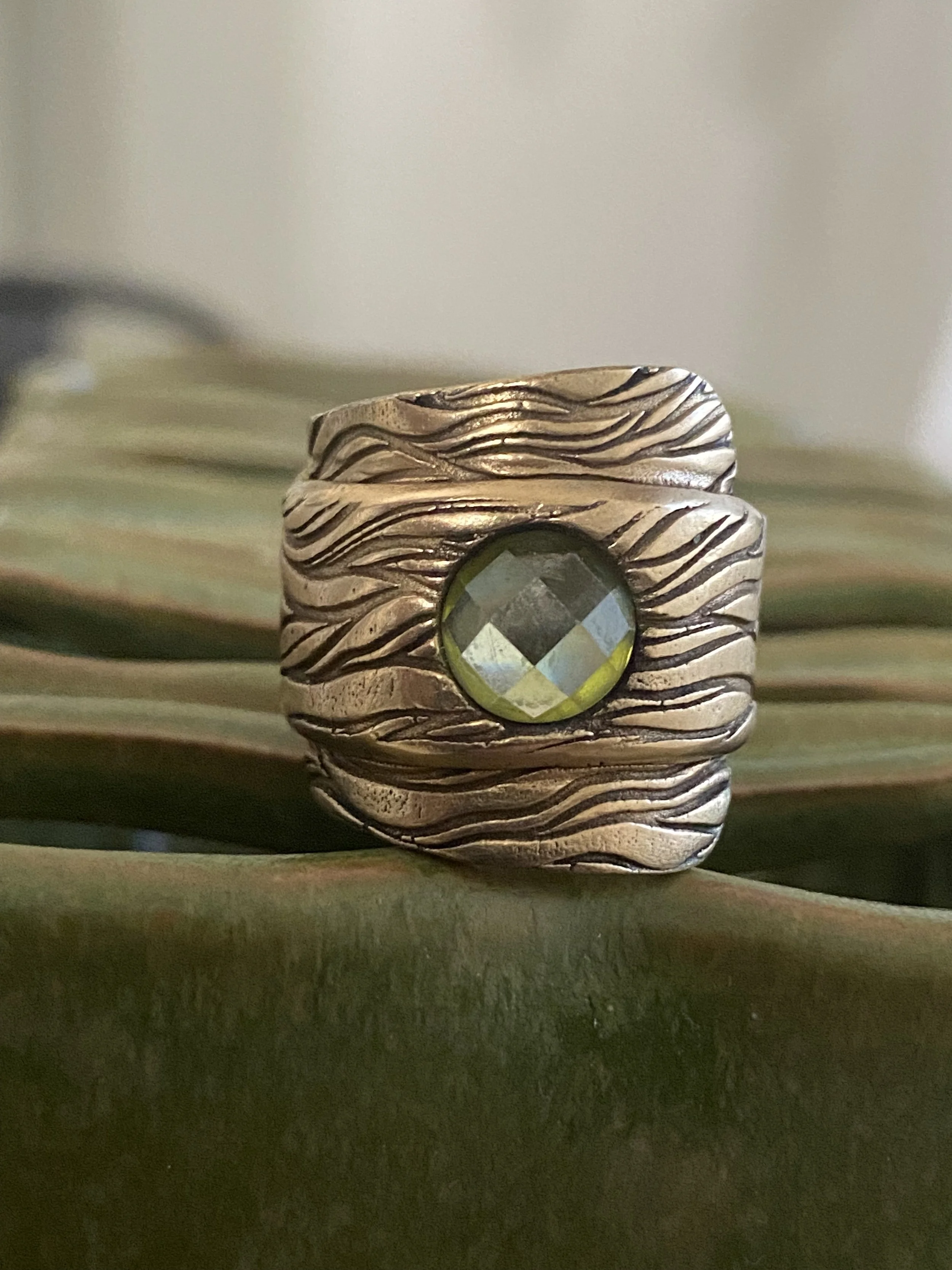 Close-up of a silver ring with a small green gemstone, featuring a textured, wavy design.