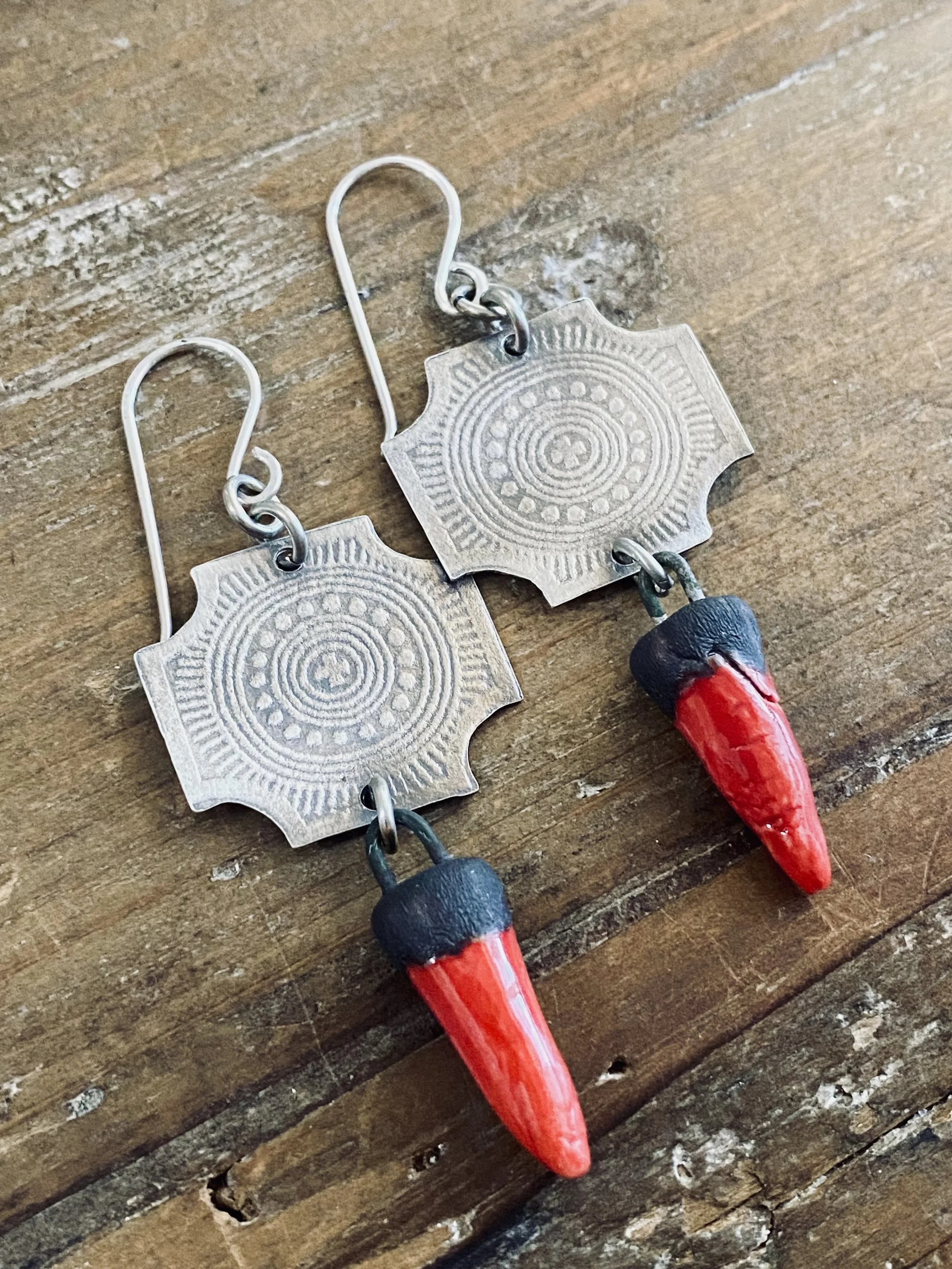 Close-up of a set of handmade earrings with metal discs featuring concentric circles and patterns, hanging from silver hooks, and decorated with red and black beads at the bottom, placed on a wooden surface.