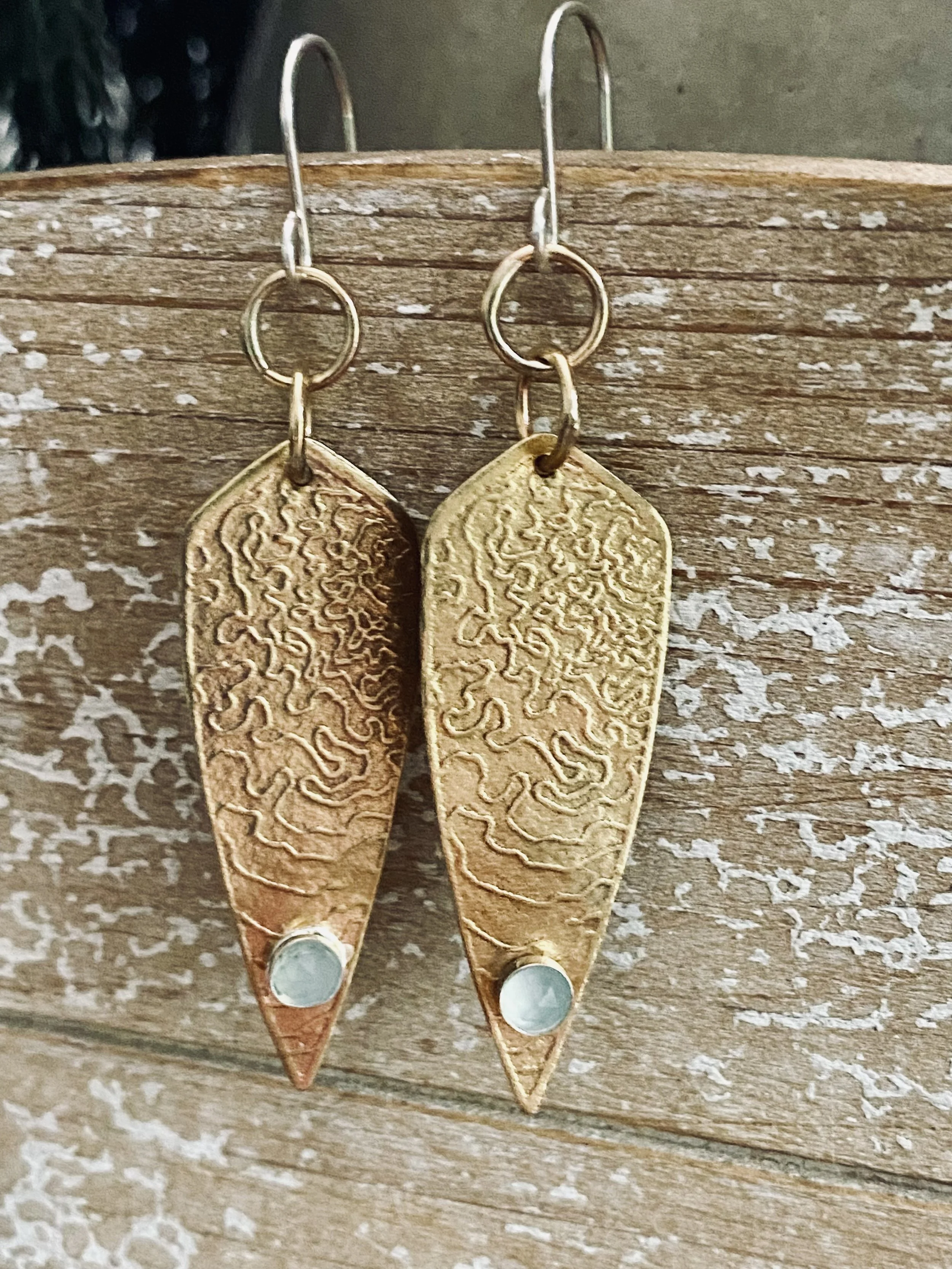 A pair of gold earrings with textured, engraved rectangular pendants hanging from small hooks. The earrings are attached to a weathered piece of wood.