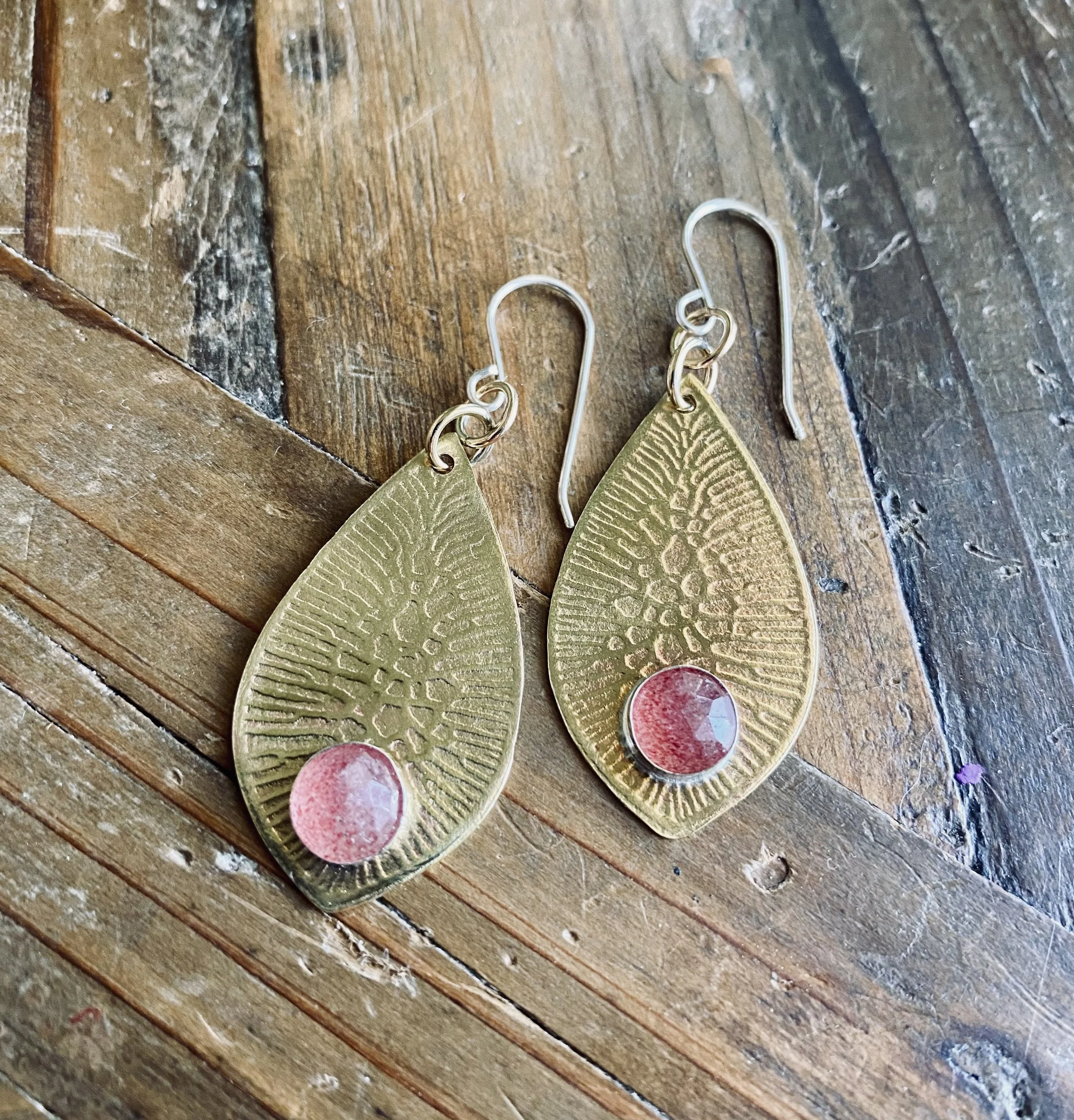 Close-up of gold-colored earrings with circular design and red accents, placed on a wooden surface with visible grain and cracks.