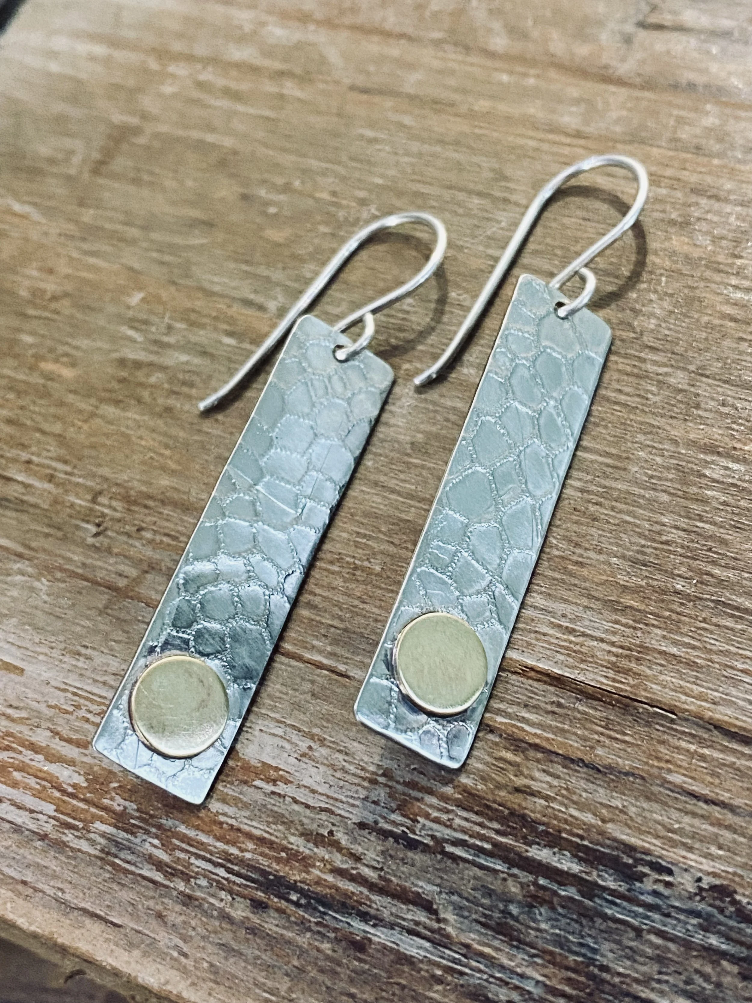 Close-up of a pair of silver earrings with rectangular textured pendants and circular elements at the bottom, lying on a wooden surface.