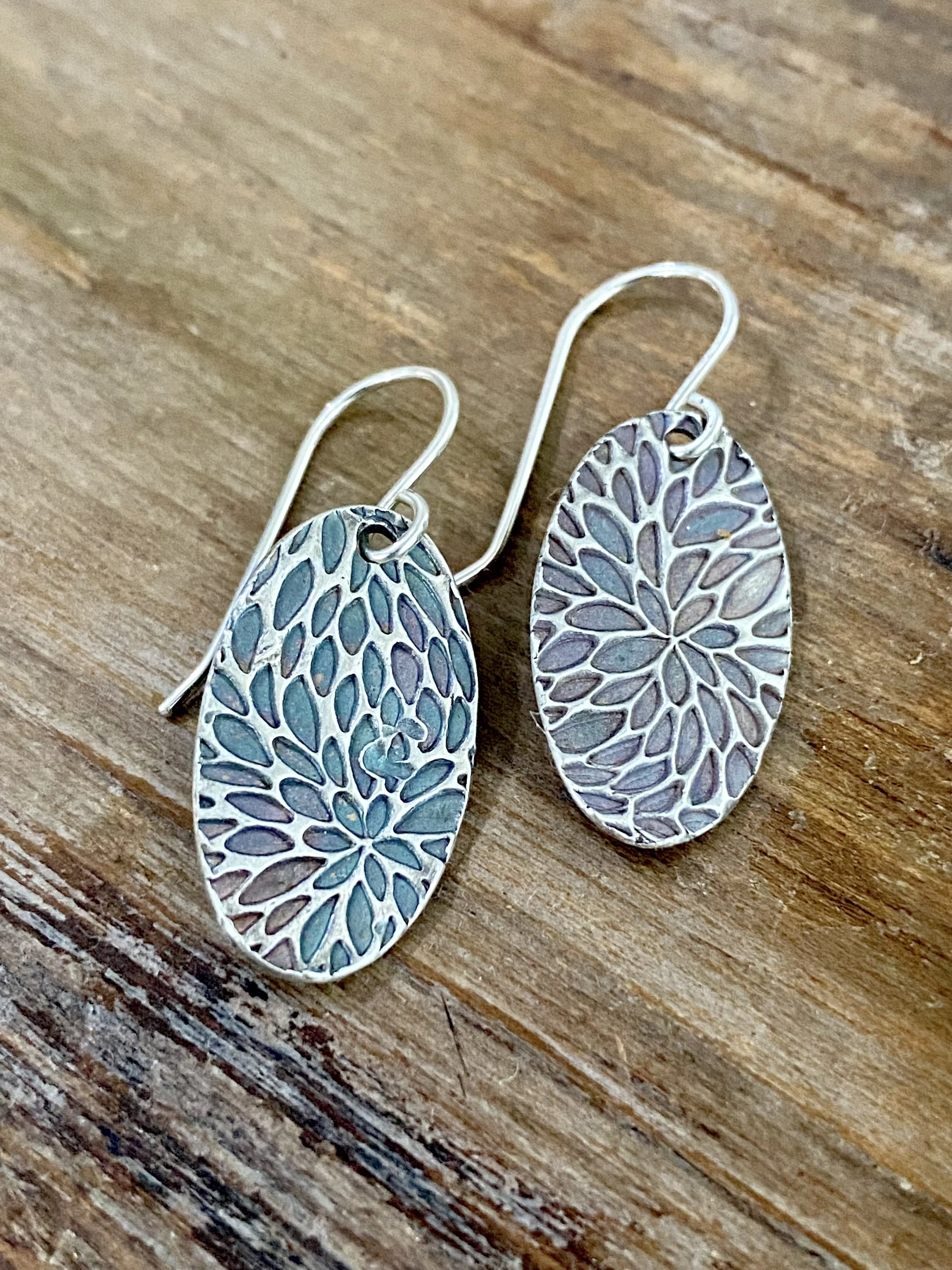 Colorful oval earrings with leaf-like pattern resting on a textured wooden surface.