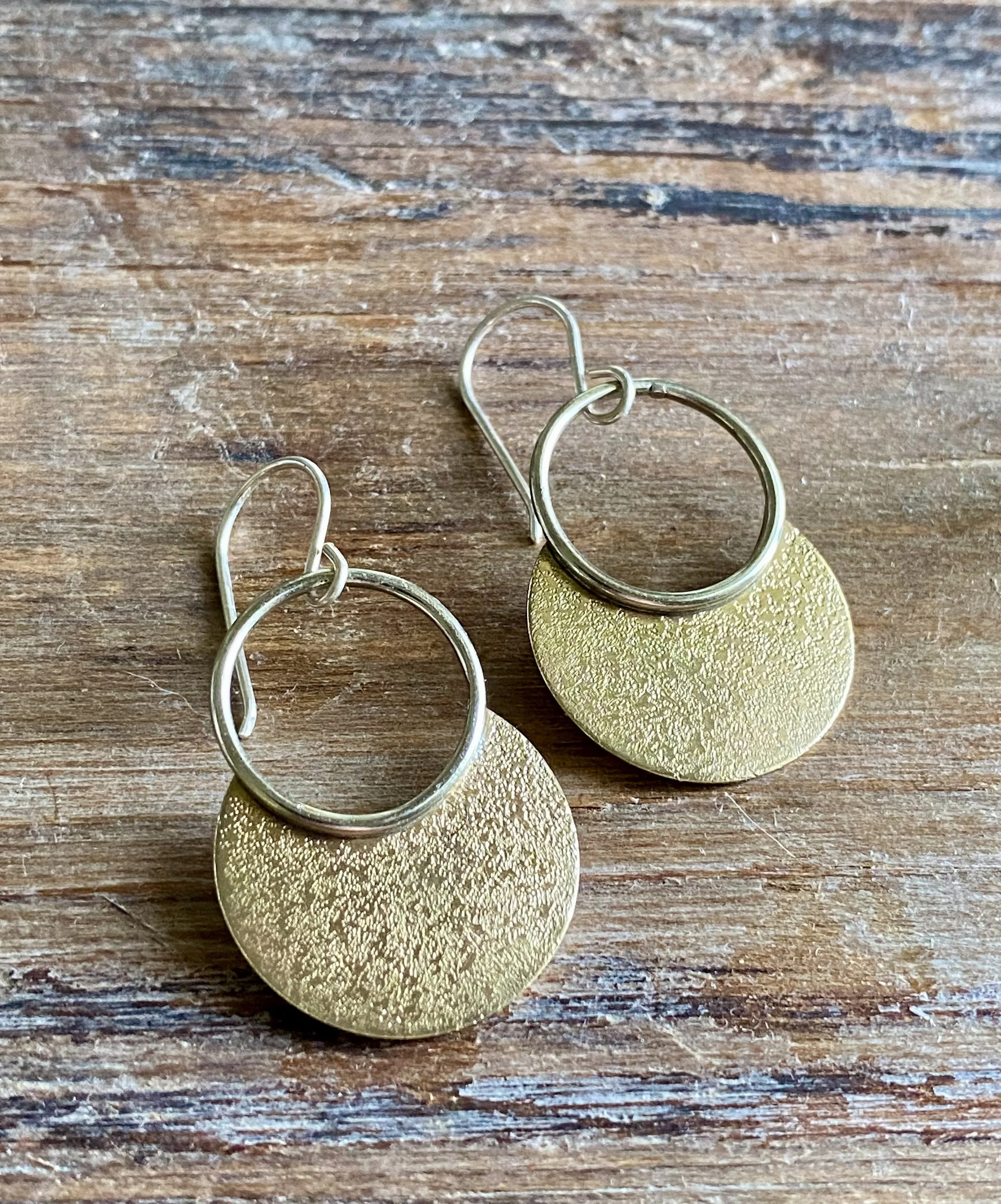 Gold hoop earrings with round textured disks hanging from the hoops, placed on a wooden surface.