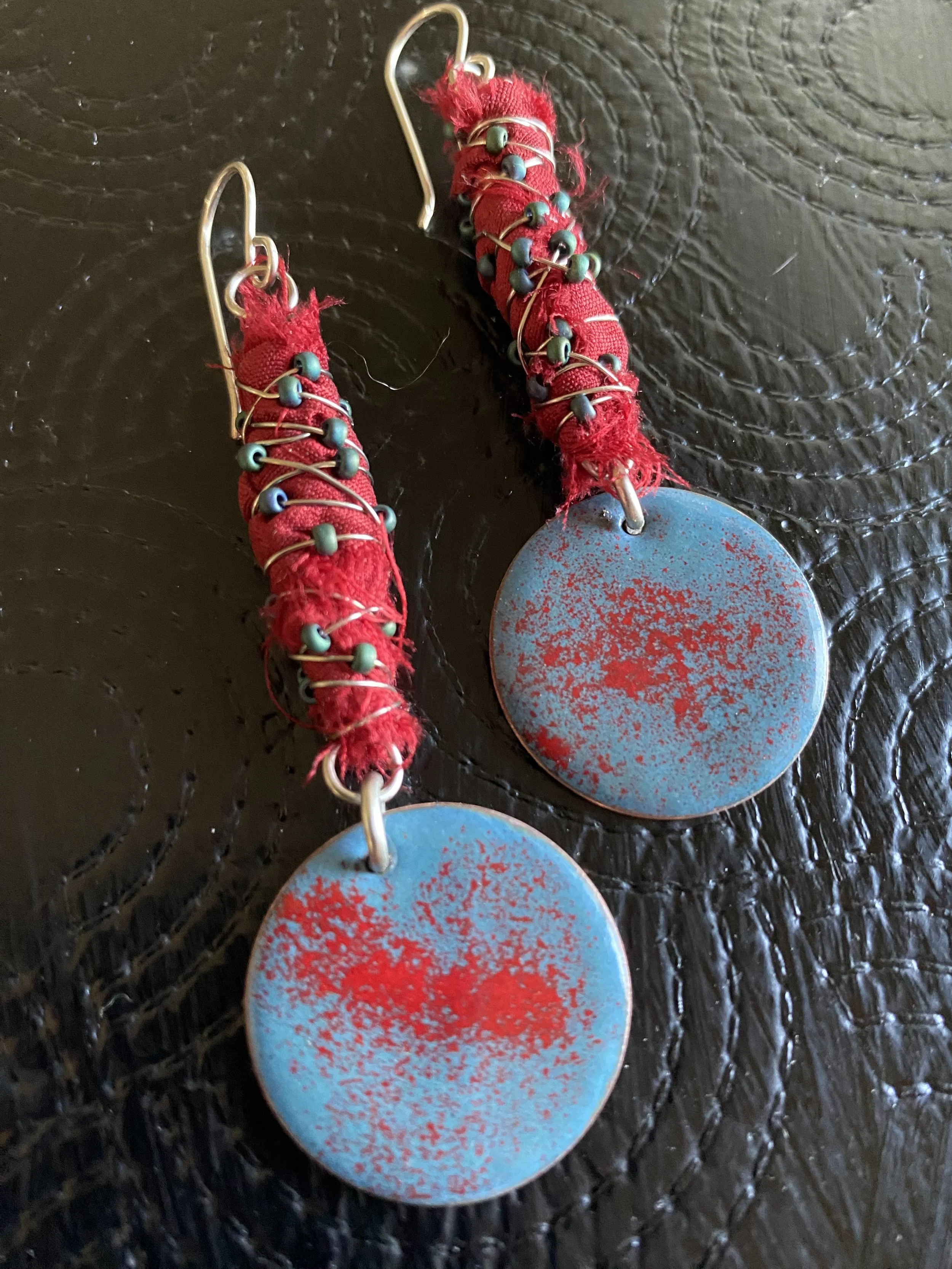 A pair of earrings with blue circular discs, wrapped with red fabric and silver wire, lying on a black textured surface.