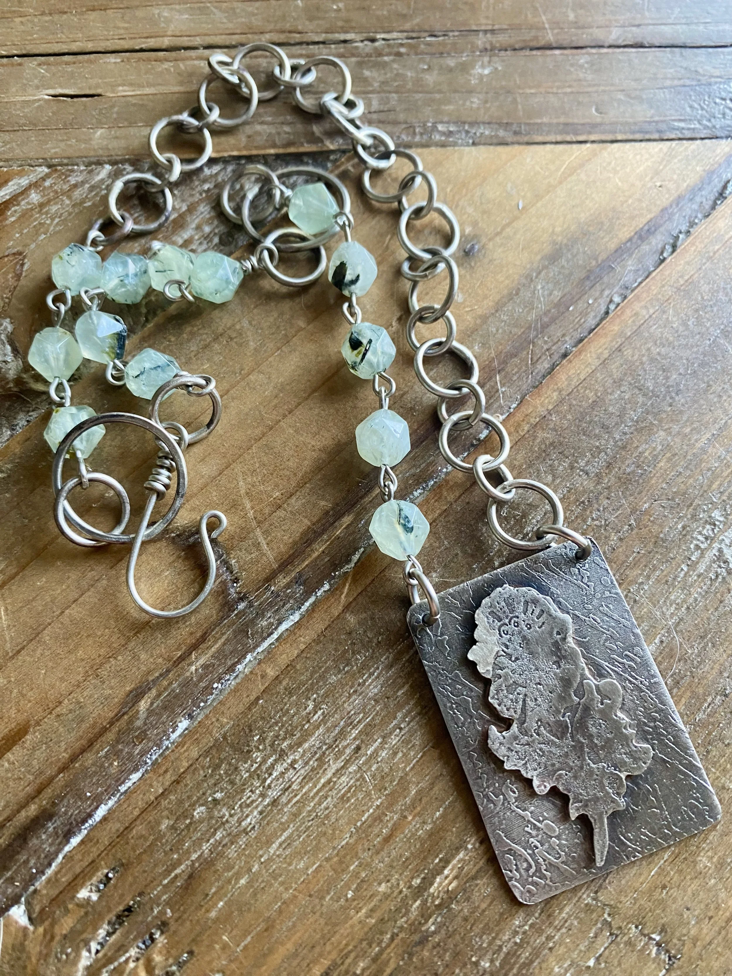 A silver jewelry chain with light green beads and a large, vintage-style metal pendant on a wooden surface.