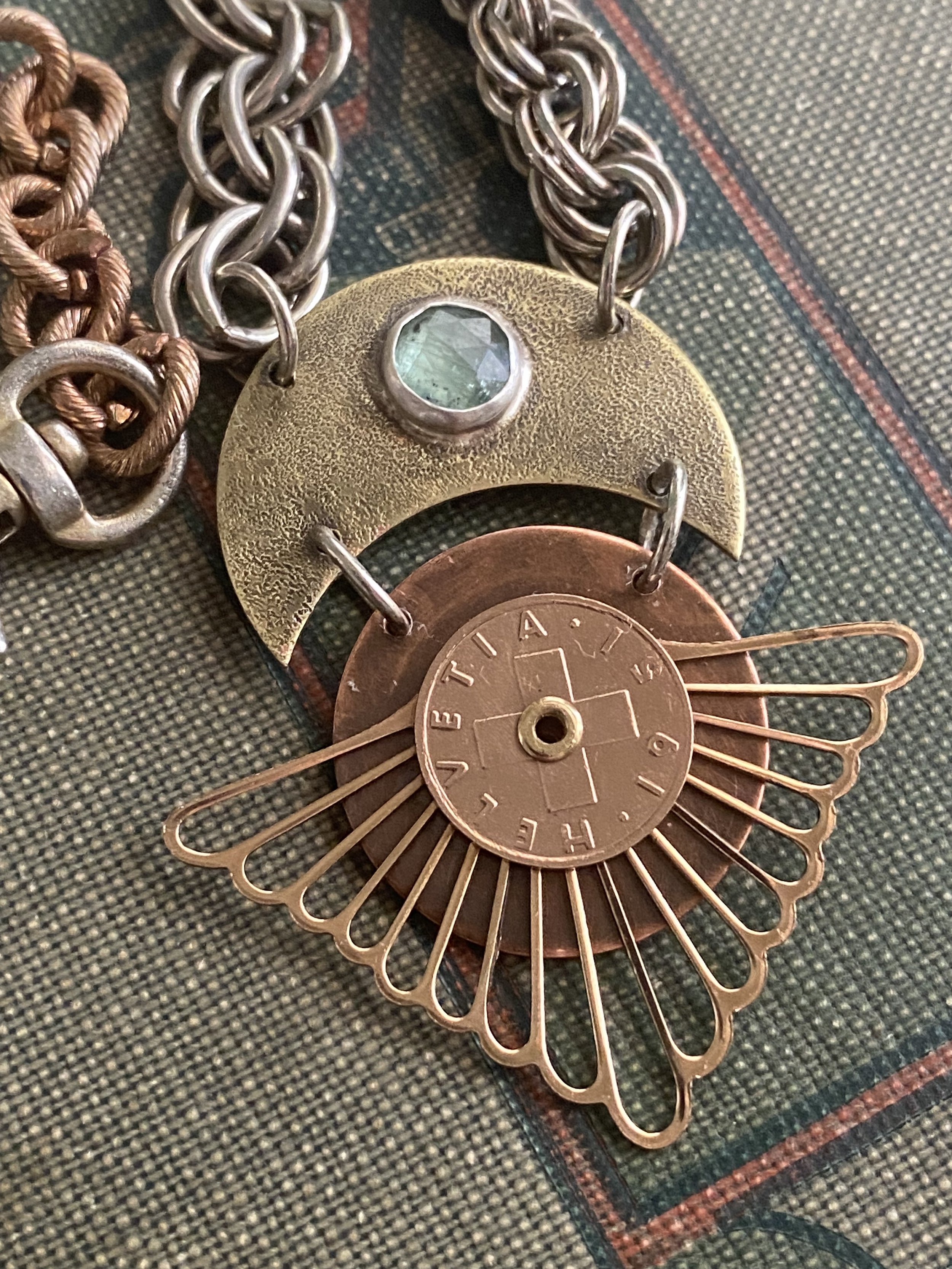 Close-up of layered metallic jewelry, including chains and a decorative piece with a gemstone and a coin with a compass design, lying on textured fabric.