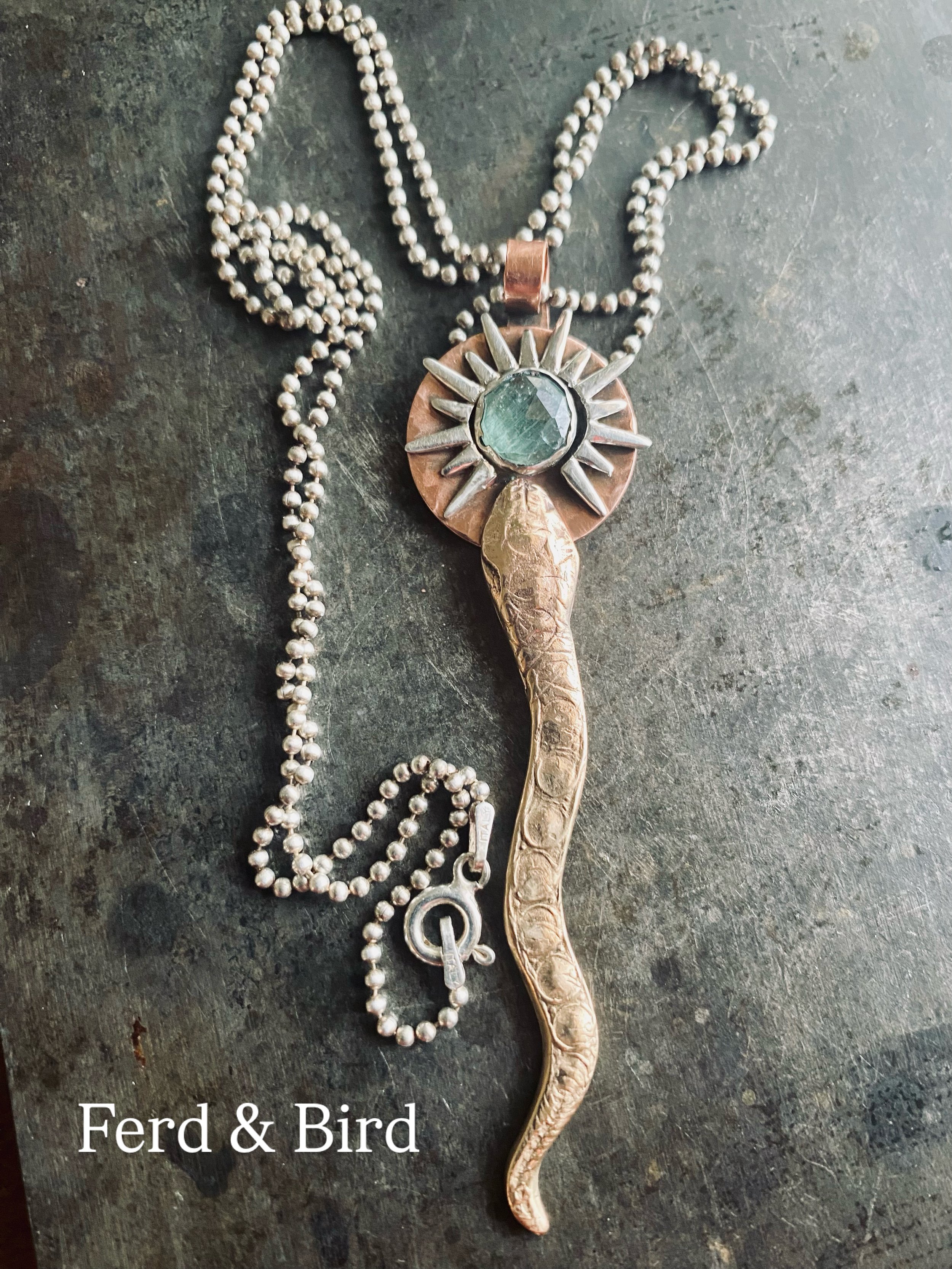 A necklace made of small pearl-like beads with a pendant that features a sun design and a large green gemstone in the center, placed on a dark textured surface.