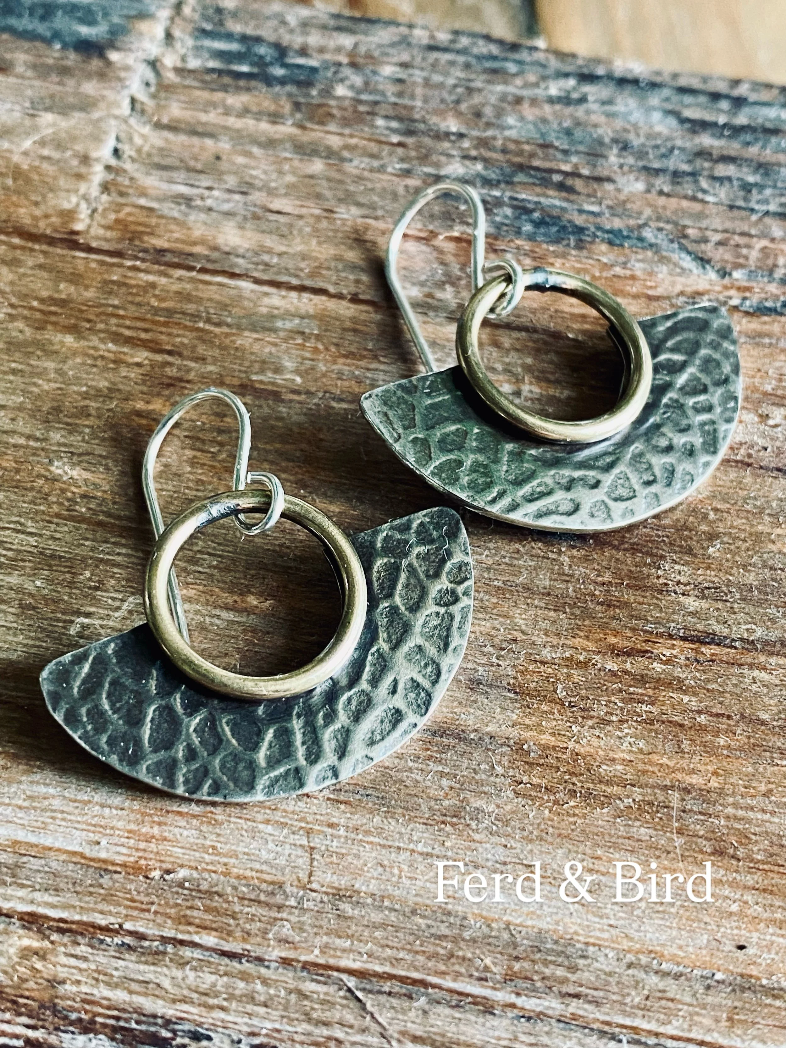 Close-up of a pair of handmade earrings with textured metallic leaves and heart-shaped hooks, resting on a rustic wooden surface