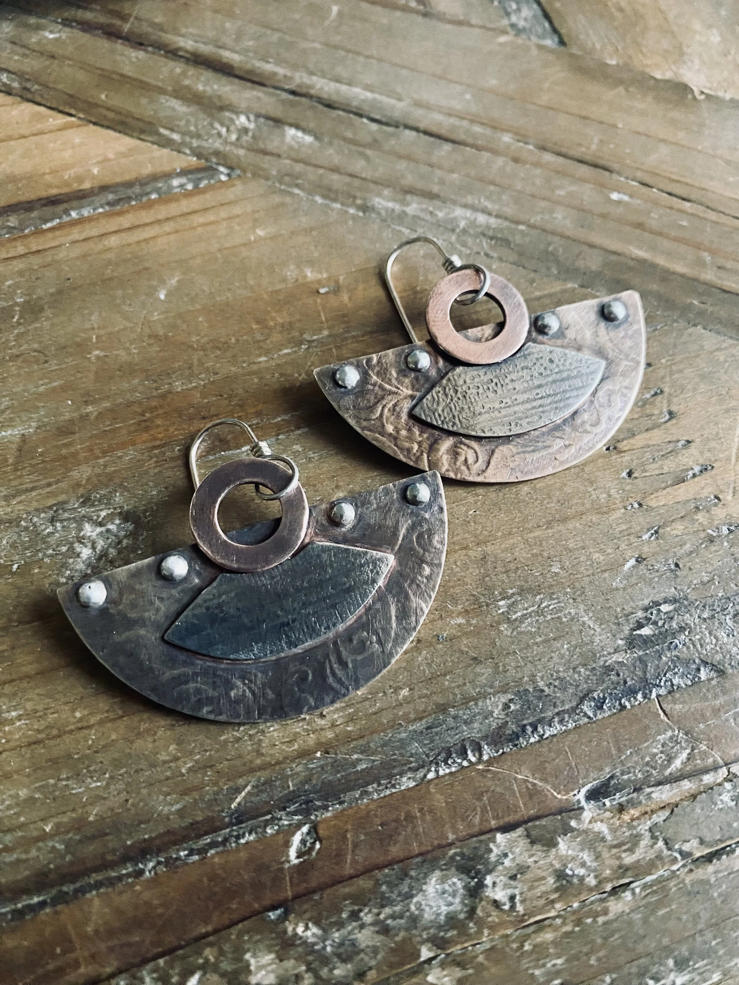 Close-up of a pair of dangling earrings with round and semi-circular metal pieces on a textured wooden surface.