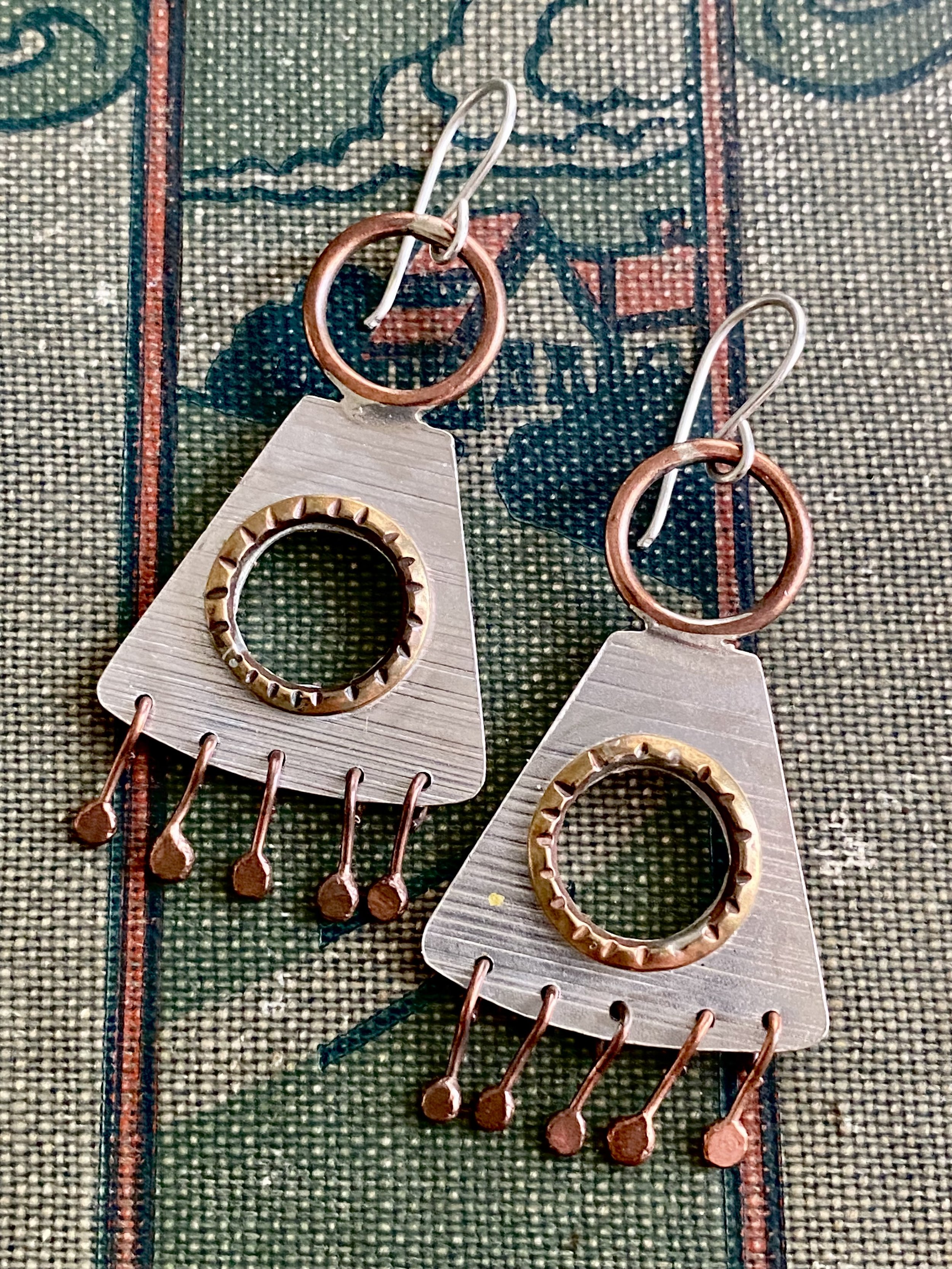 Close-up of earrings with silver bases, bronze rings, and small bronze charms, placed on fabric with a colorful, patterned design.