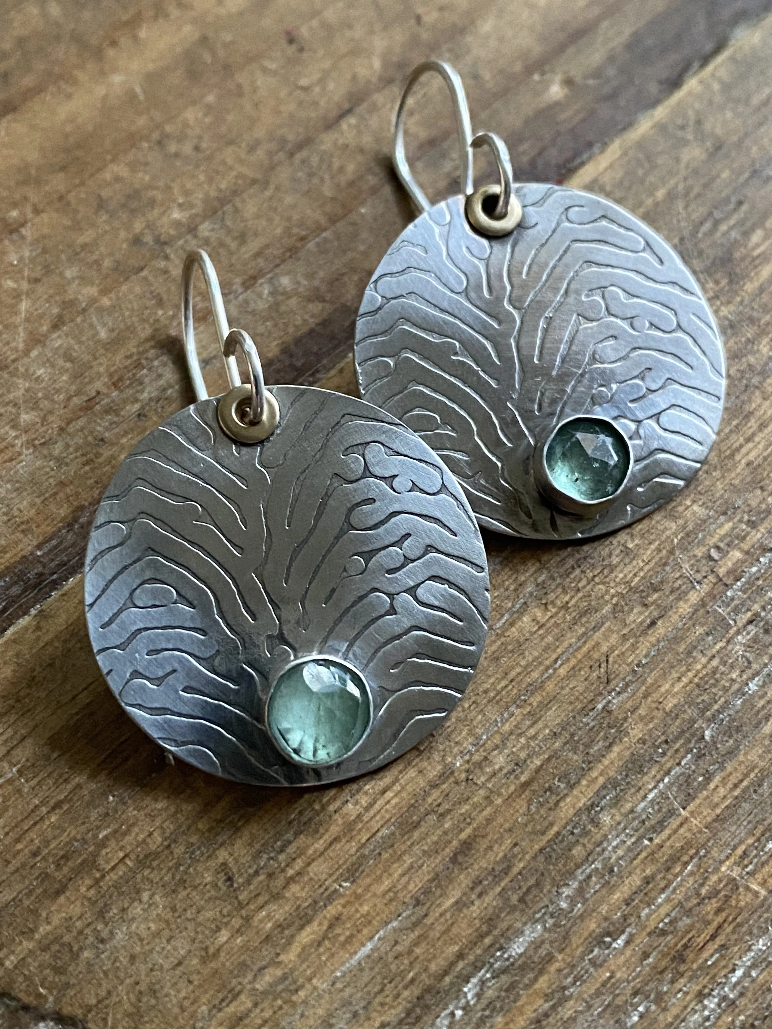 A pair of silver earrings with a tree pattern and a small green gemstone at the bottom, resting on a wooden surface.