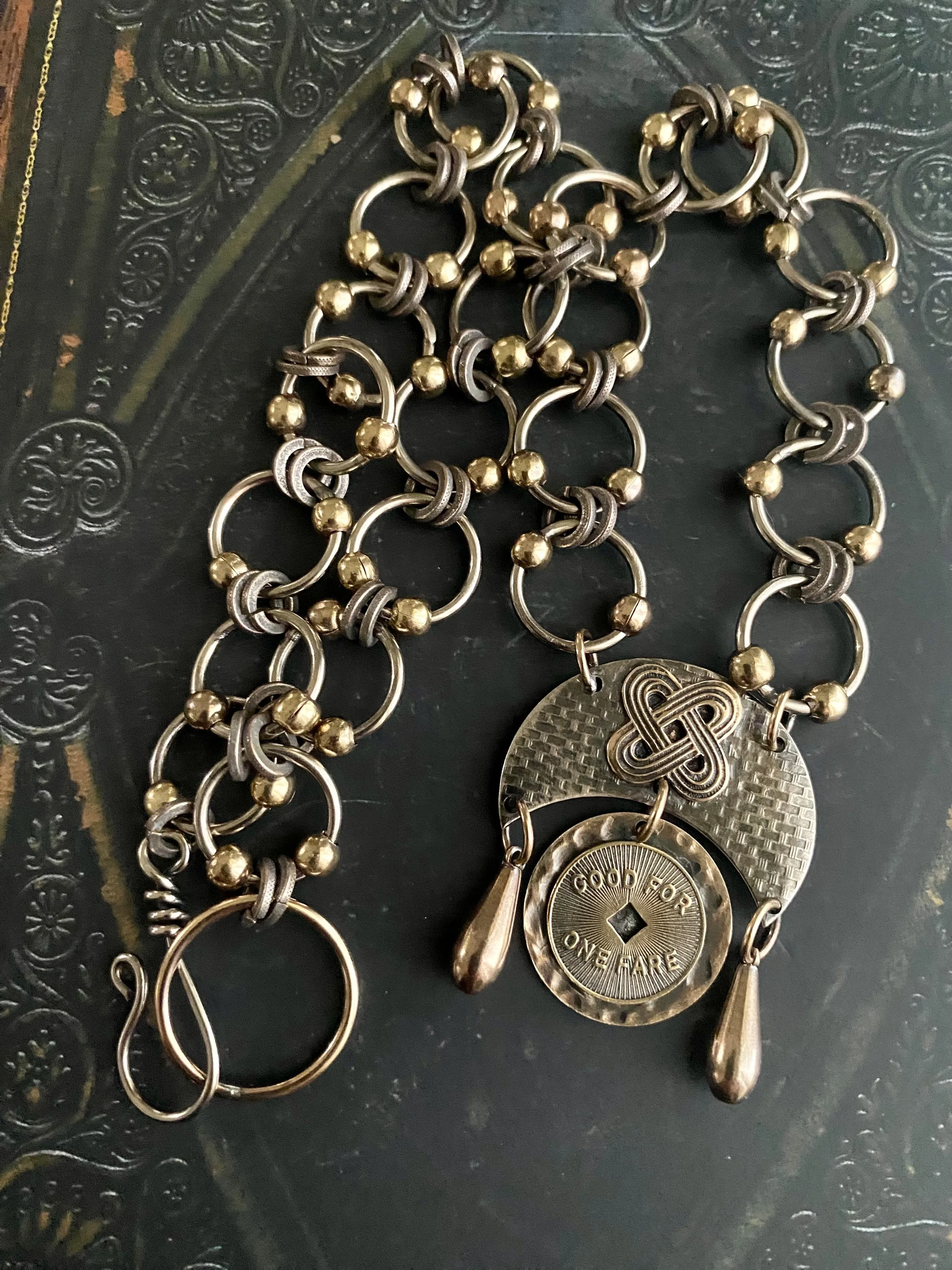 Assorted vintage jewelry including layered bracelets with gold and silver beads, a large round coin charm engraved with "Good for one fare," and various metal components, laid out on a decorative dark surface.