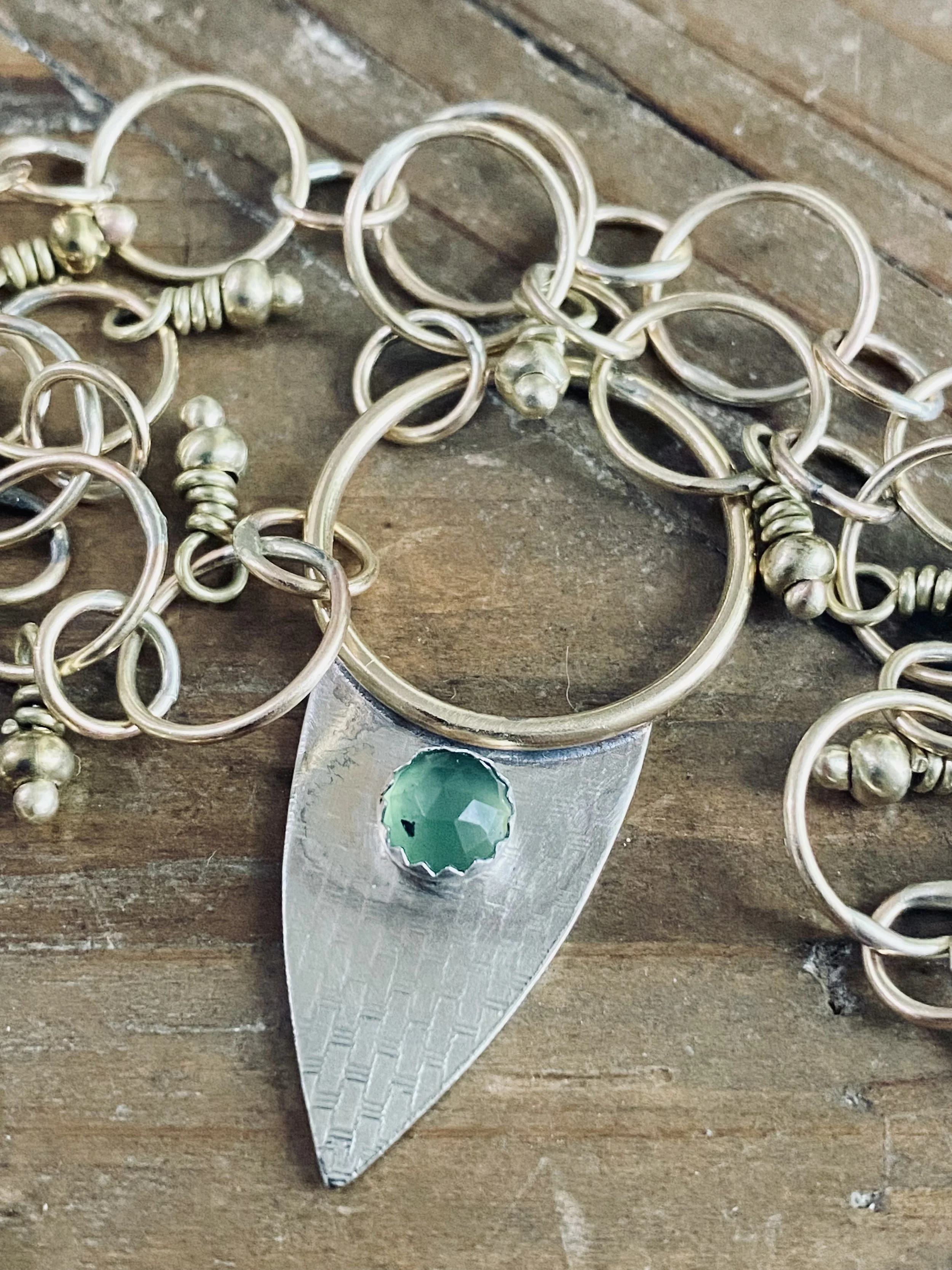 Silver jewelry with circular and spiral designs, including a piece with a large green gemstone, displayed on a wooden surface.