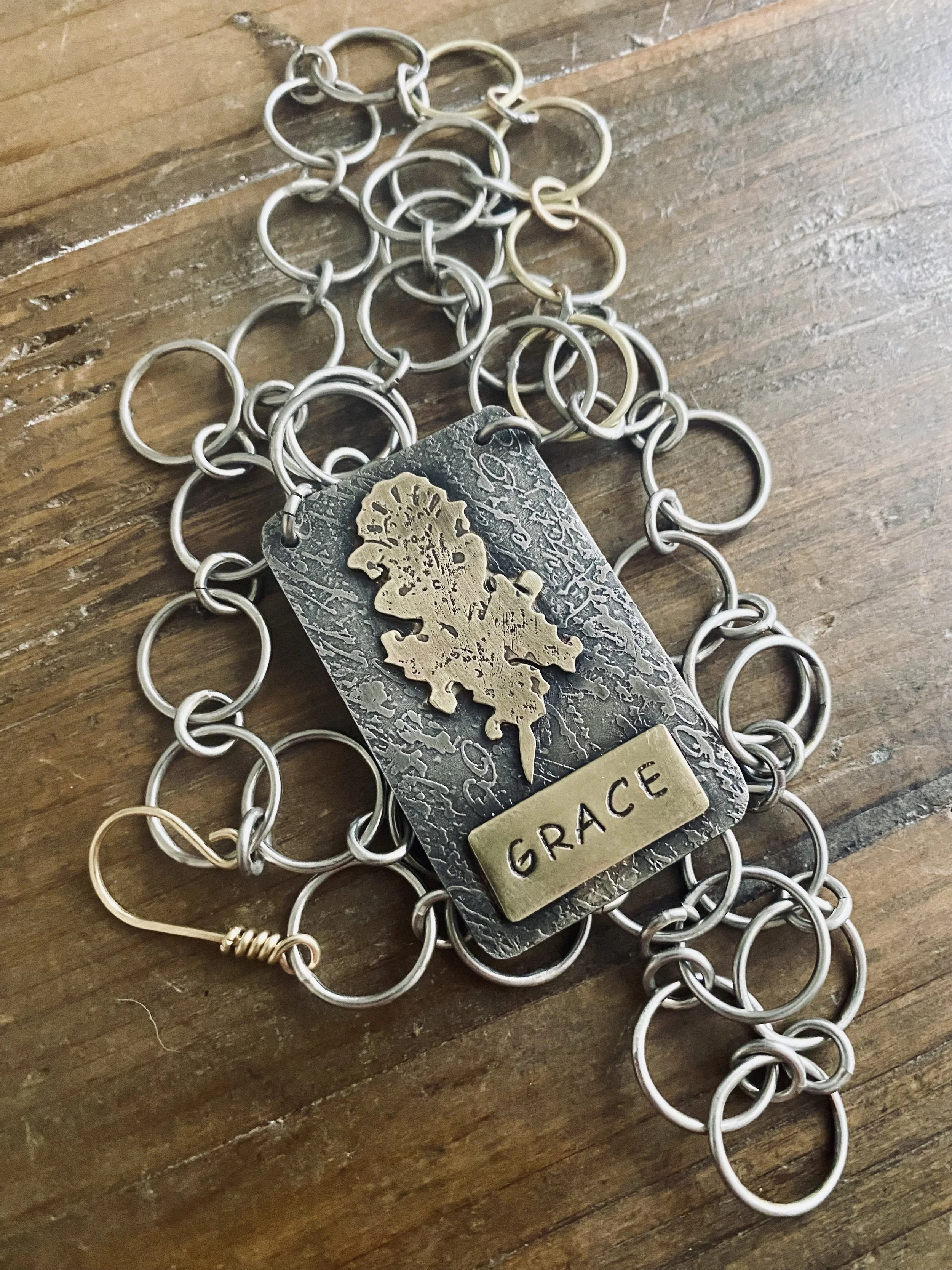 Silver metal chain with a rectangular metal tag reading 'GRACE' and a relief map of the world, lying on a wooden surface.