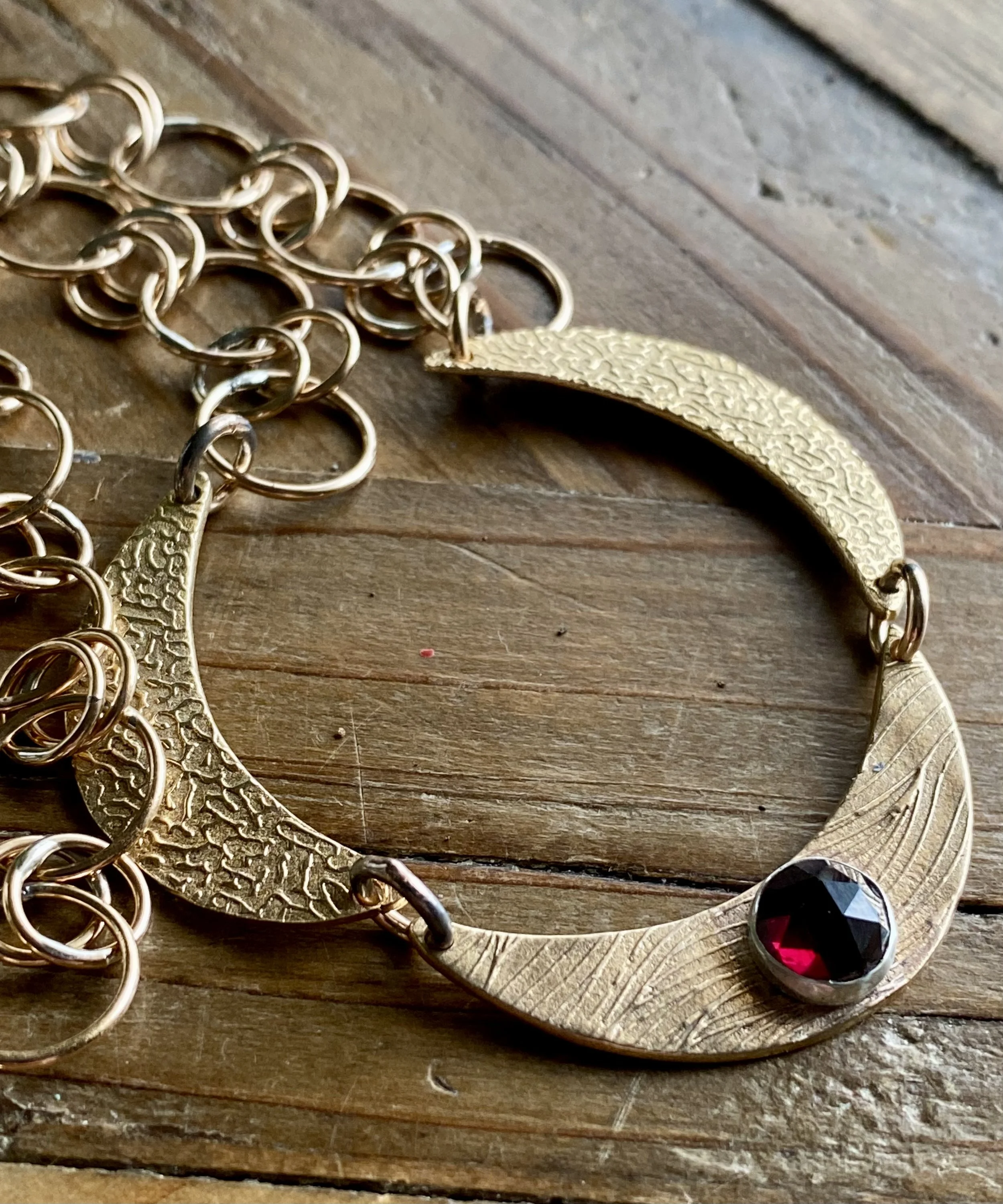 Close-up of gold-colored jewelry with a textured crescent-shaped pendant, intertwined with gold rings, on a wooden surface, and a person wearing black shoes with red soles visible at the bottom.