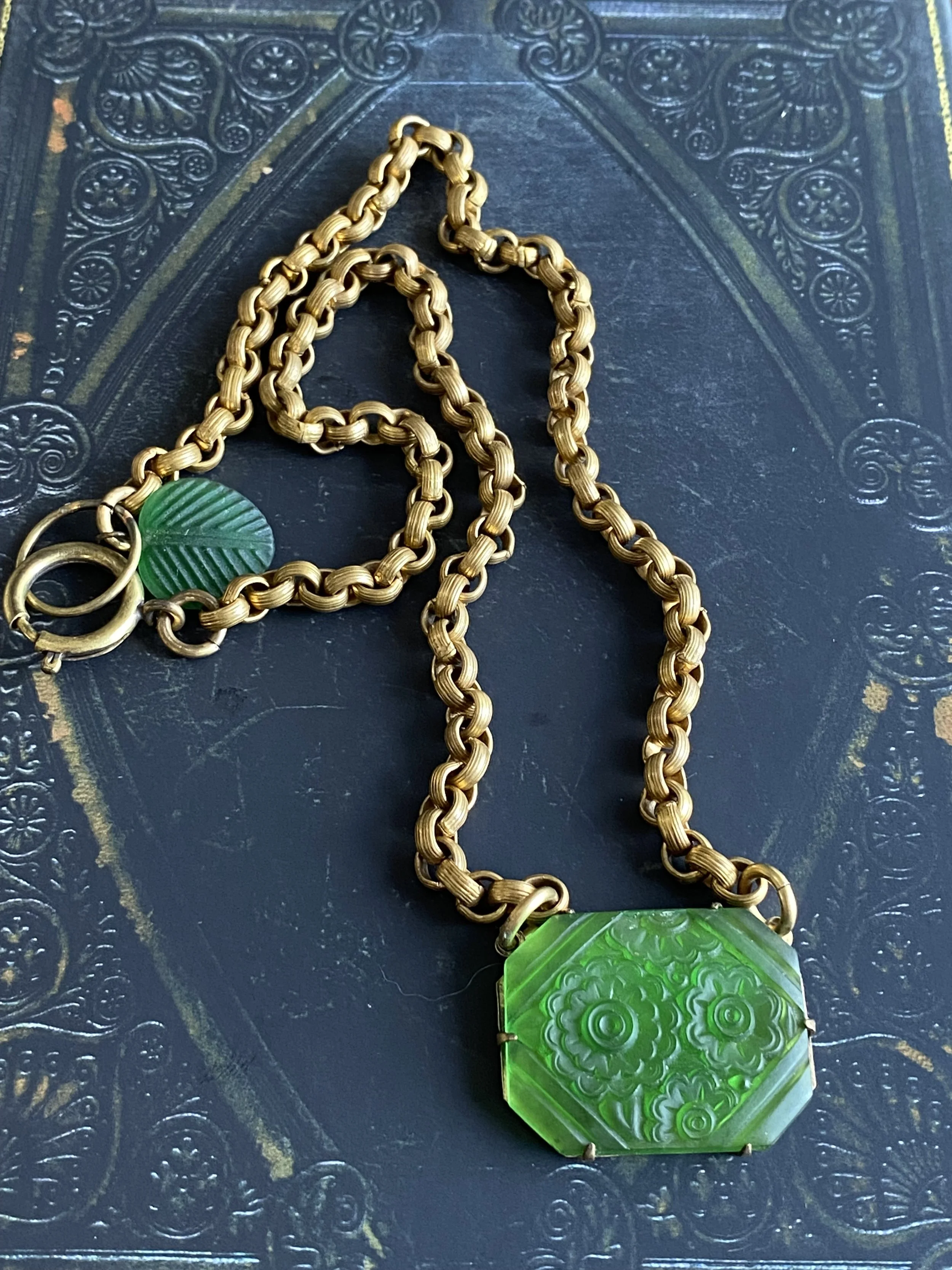 A gold chain necklace with a large green carved jade pendant and a smaller green carved jade detail on the clasp, resting on a textured dark surface.
