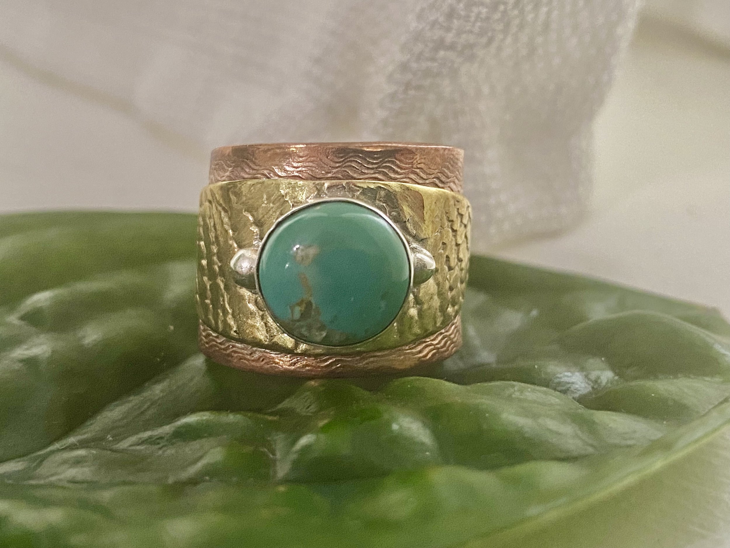 A close-up of two rings, one with a large turquoise stone and the other made of textured metal, placed on a green leaf with a soft, light background.