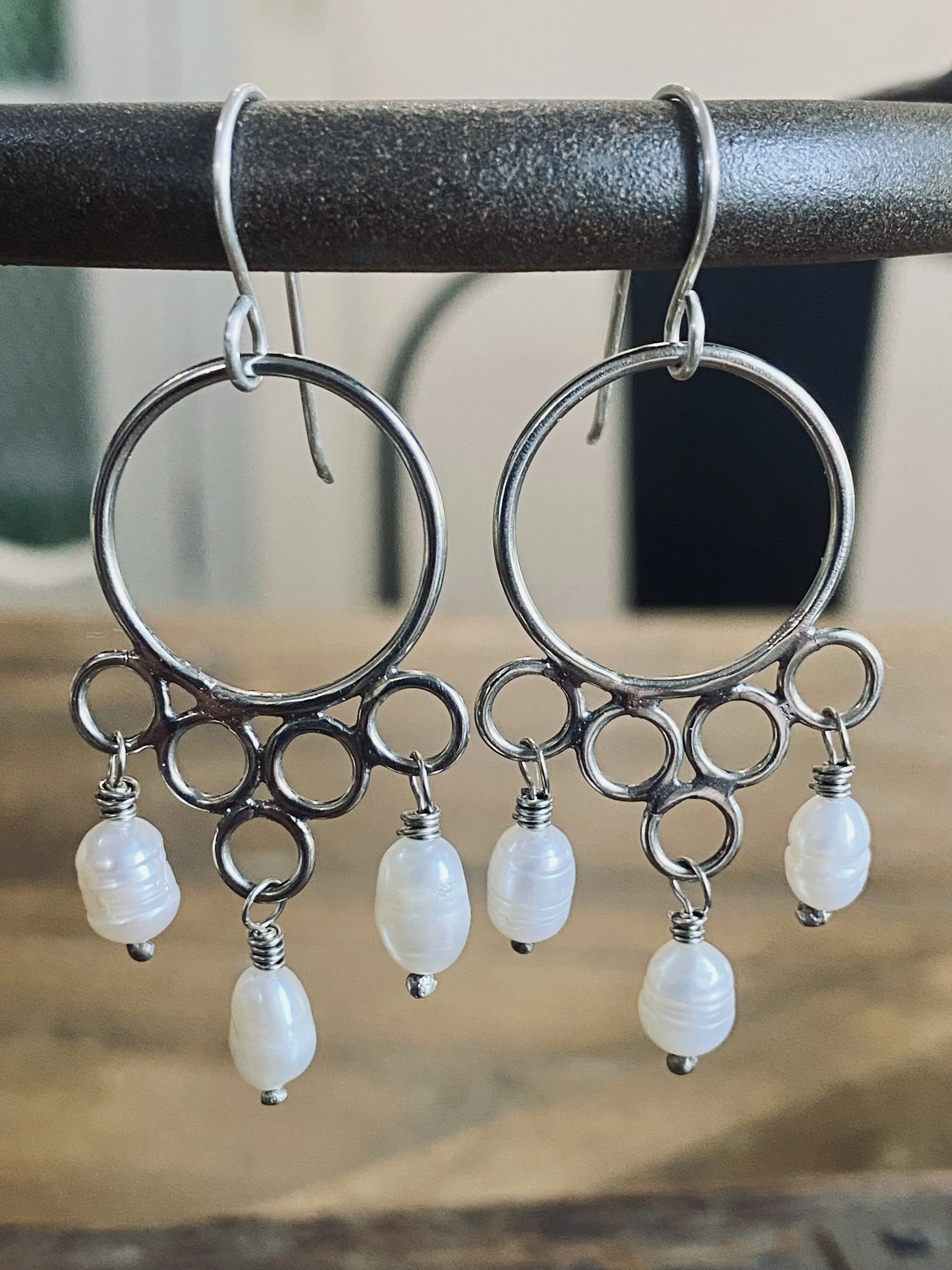 Pair of dangling earrings with oval-shaped pearl-like beads hanging from a metal framework with loop designs, hanging from a black rail.