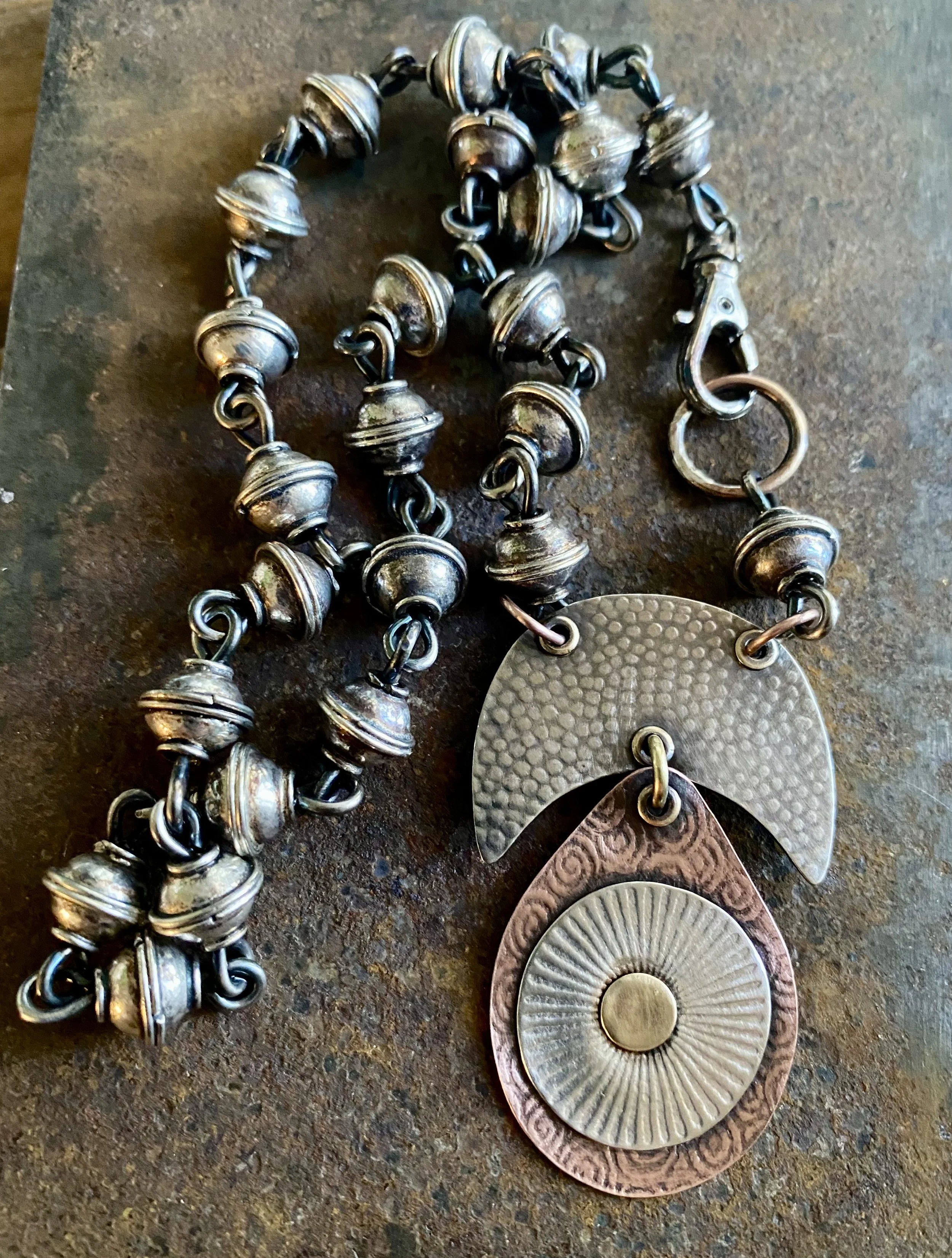 Close-up of a metallic jewelry chain with large round beads and pendants, placed on a rough surface. The pendants are round, textured, and decorated with patterns.