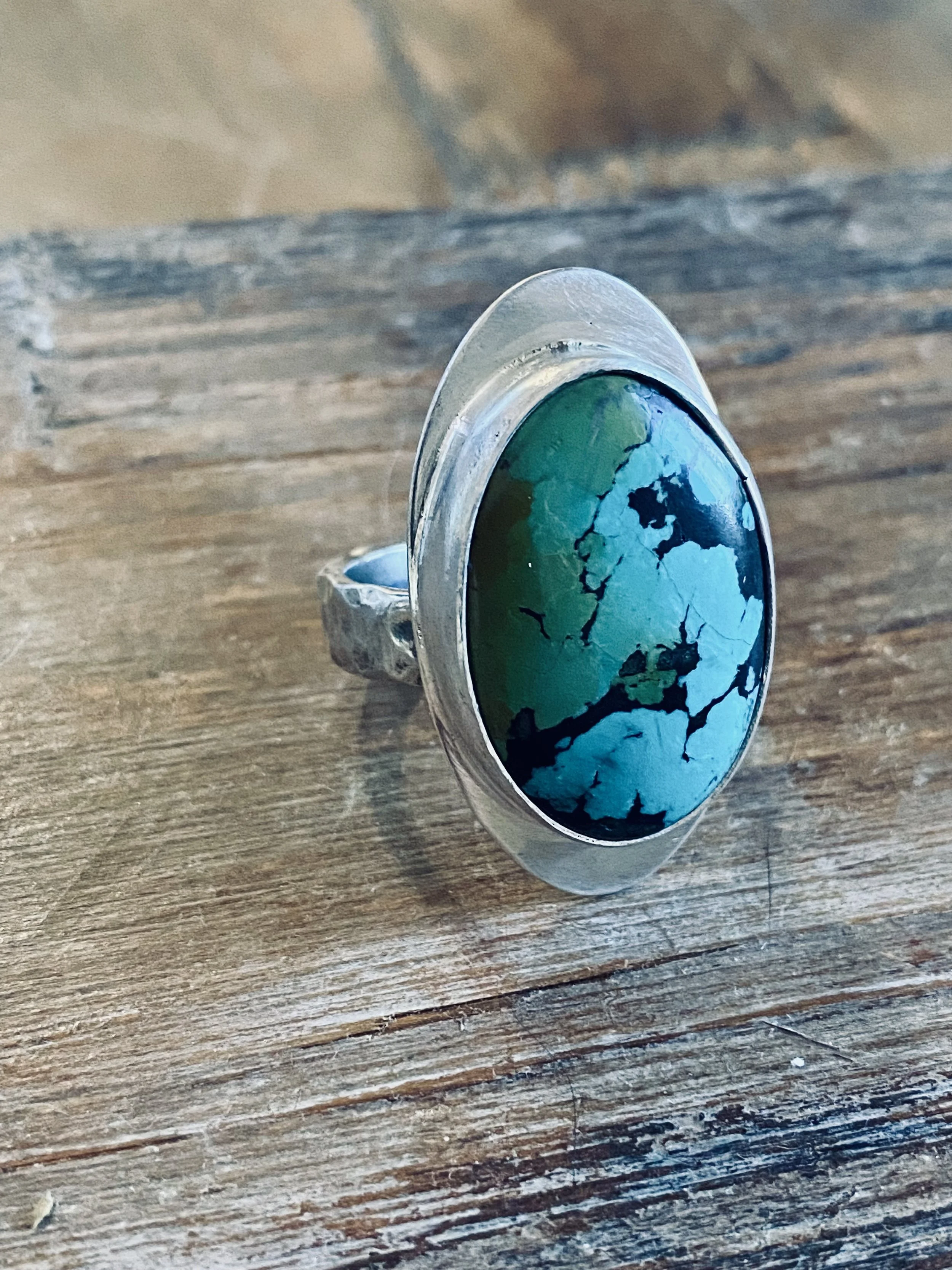 A silver ring with a large oval turquoise stone featuring black veining, placed on a wooden surface.