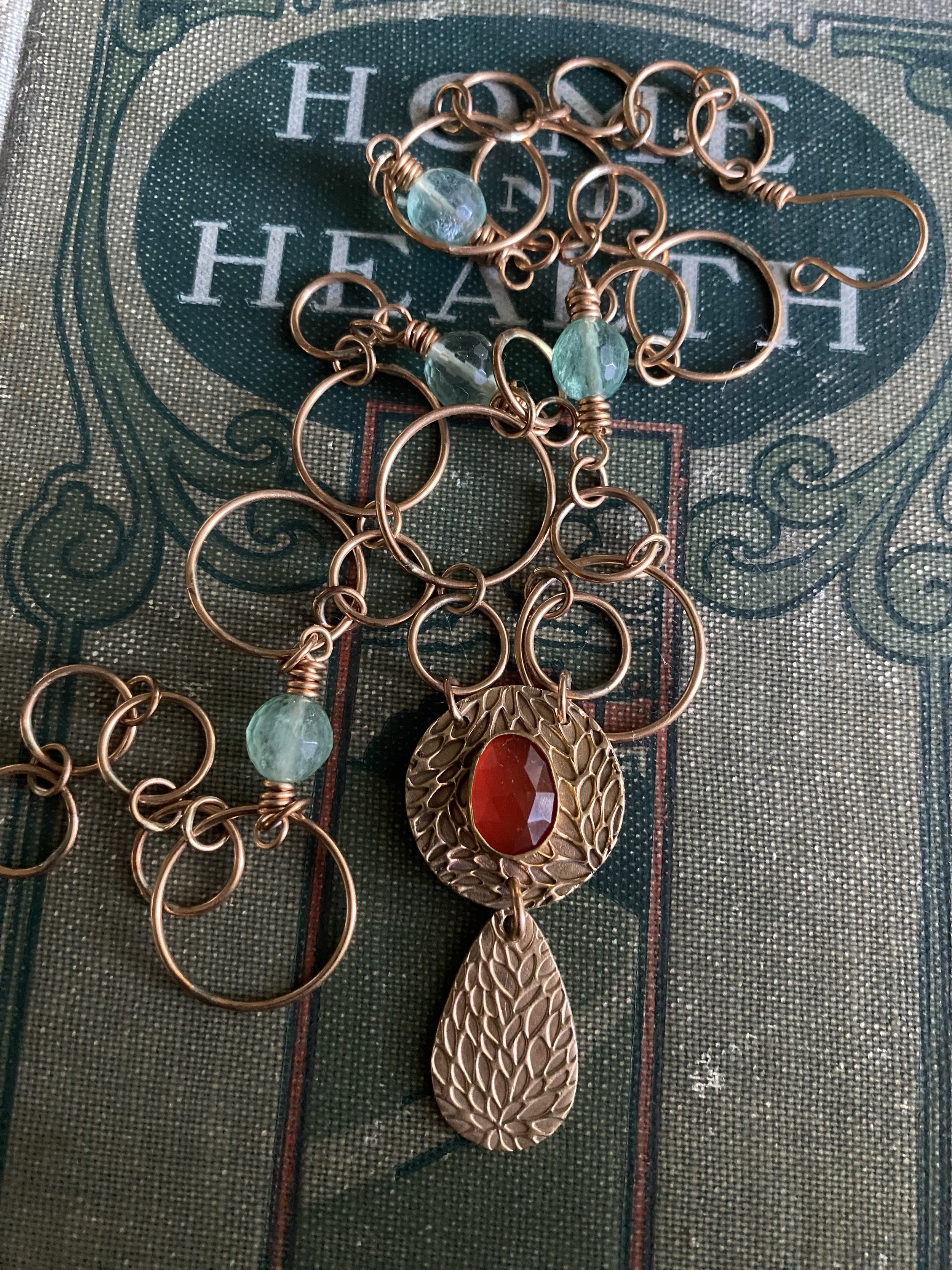 Close-up of a jewelry piece with green beads, a red stone, and a dangling leaf-shaped ornament, resting on a fabric surface with the words 'The Heart' written on it.