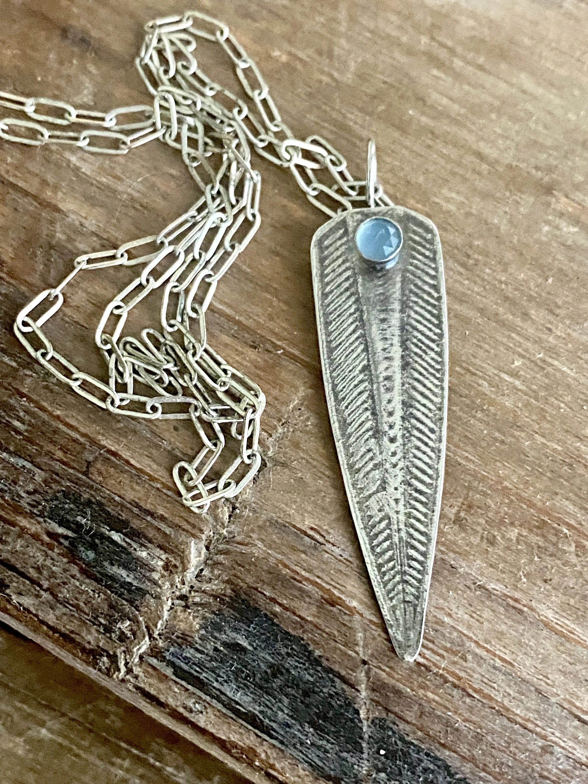 A metal chain necklace with a vintage silver feather-shaped pendant featuring a small blue gemstone embedded near the top, laid on a wooden surface.