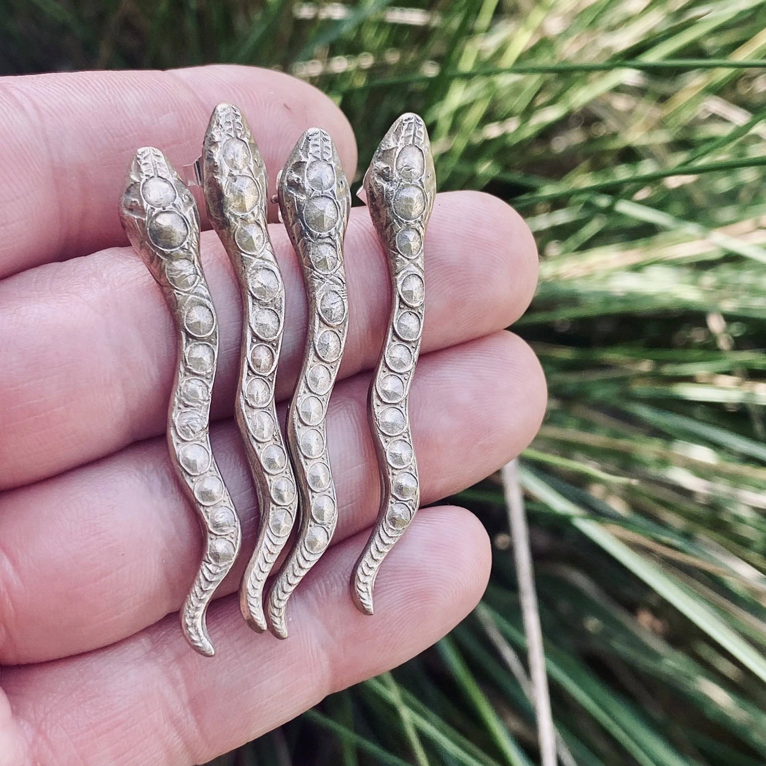 Close-up of a person's fingers with four metallic rings shaped like dragon or lizard heads, set against a background of green grass.