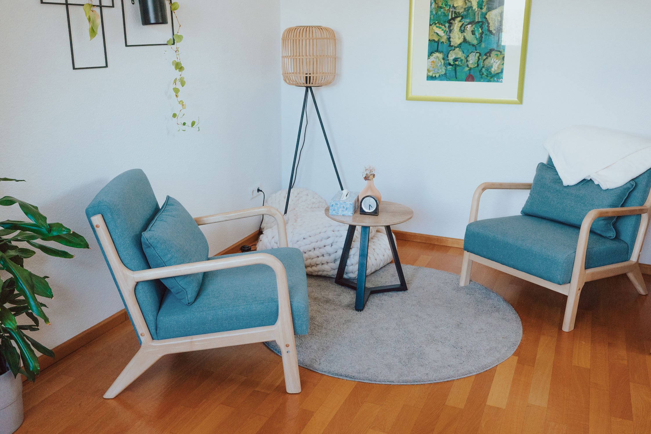 Gemütliche kleiner Praxisraum mit zwei blauen Sesseln und einem runden Holztisch mit Taschentuchbox und Deko. Im Hintergrund steht ein Stehlampe mit einem Rattan-Schirm, an der Wand hängen Kunstwerke und Pflanzen. Ein Teppich liegt auf Holzboden.
