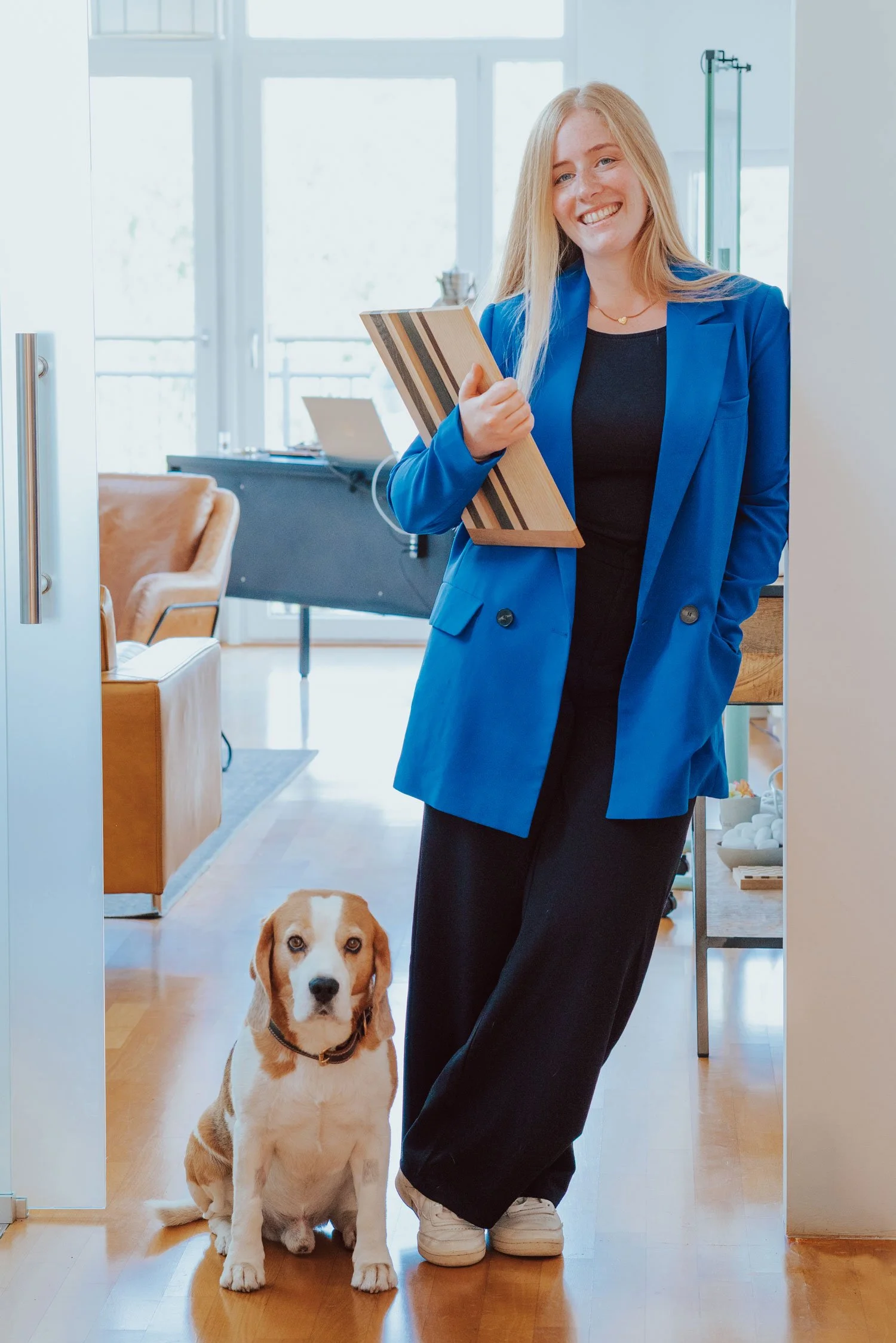 Frau Suppes in einem blauen Blazer steht am Eingang des Hauptraums der Praxis Trier. Sie hält Unterlagen und lächelt. Vor ihr sitzt Ludwig, der Praxishund mit braunen Flecken, der direkt in die Kamera schaut.